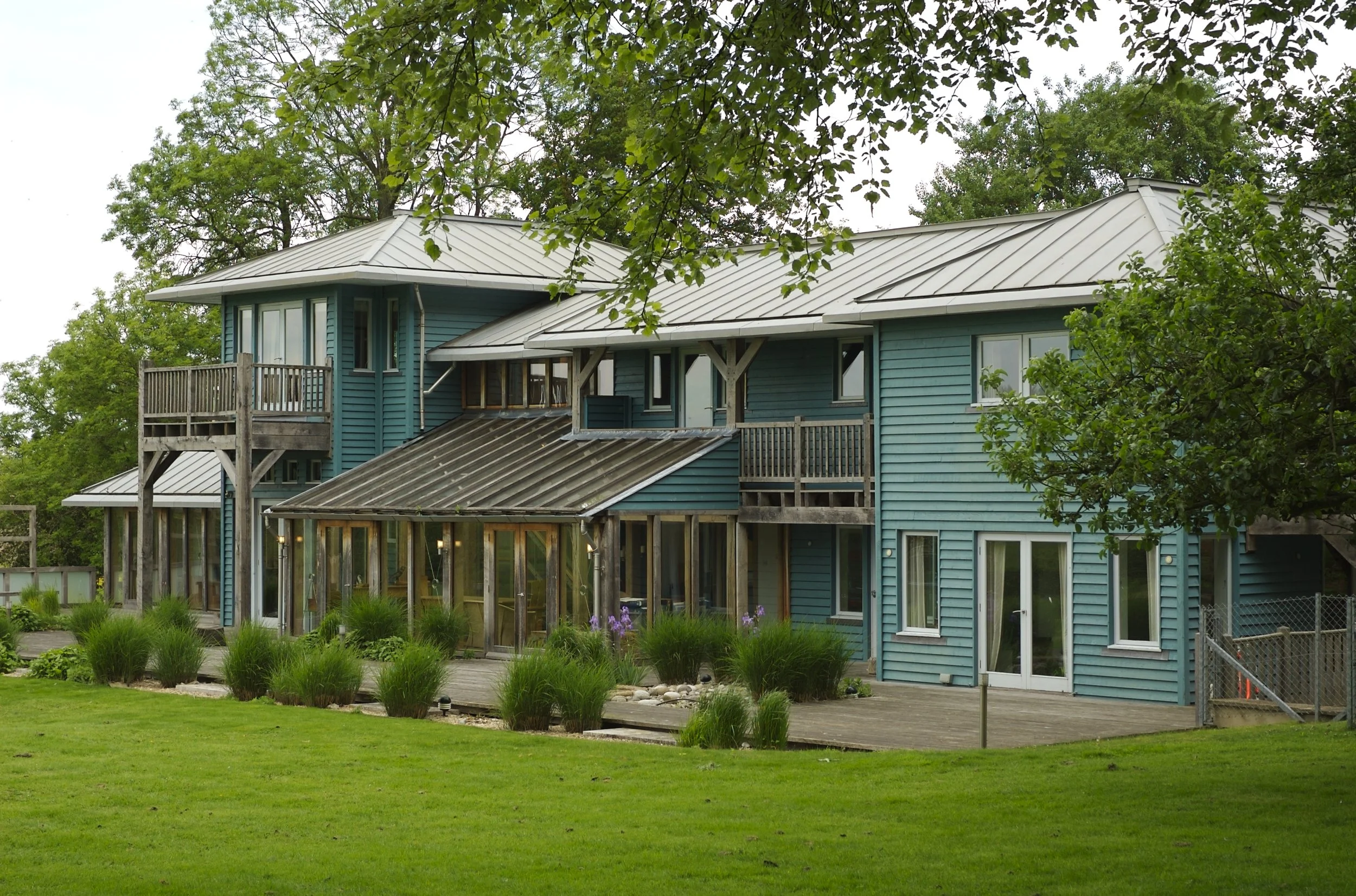 A multi-story blue house with a metal roof, balcony, and large windows, surrounded by green grass and trees.
