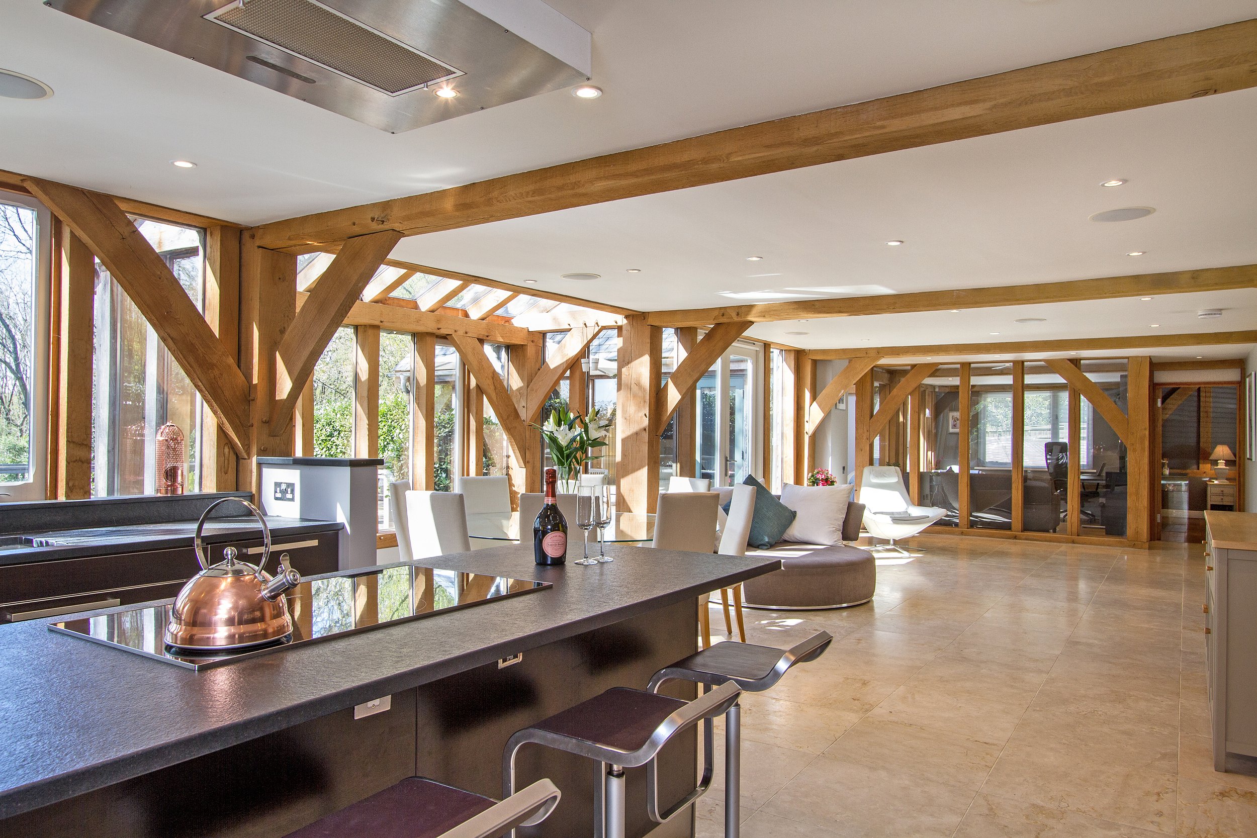 Modern open-concept living and dining area with large windows, wooden beams, and a kitchen island with a copper tea kettle and wine bottle.
