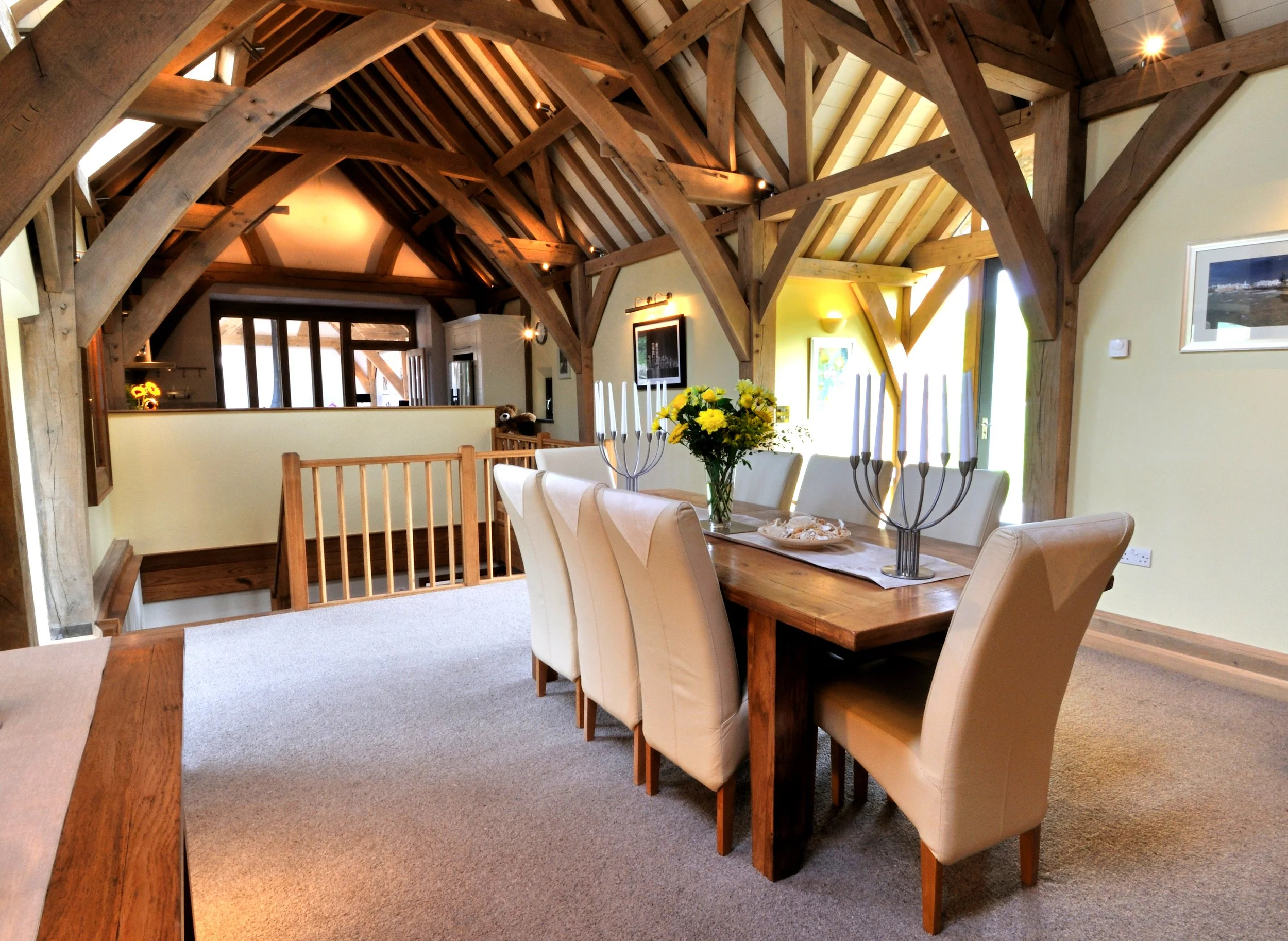 A dining room with a wooden table surrounded by beige upholstered chairs. The table is decorated with a bouquet of yellow flowers and a metal candle holder with tall white candles. The room features exposed wooden beams on the ceiling and cream-color