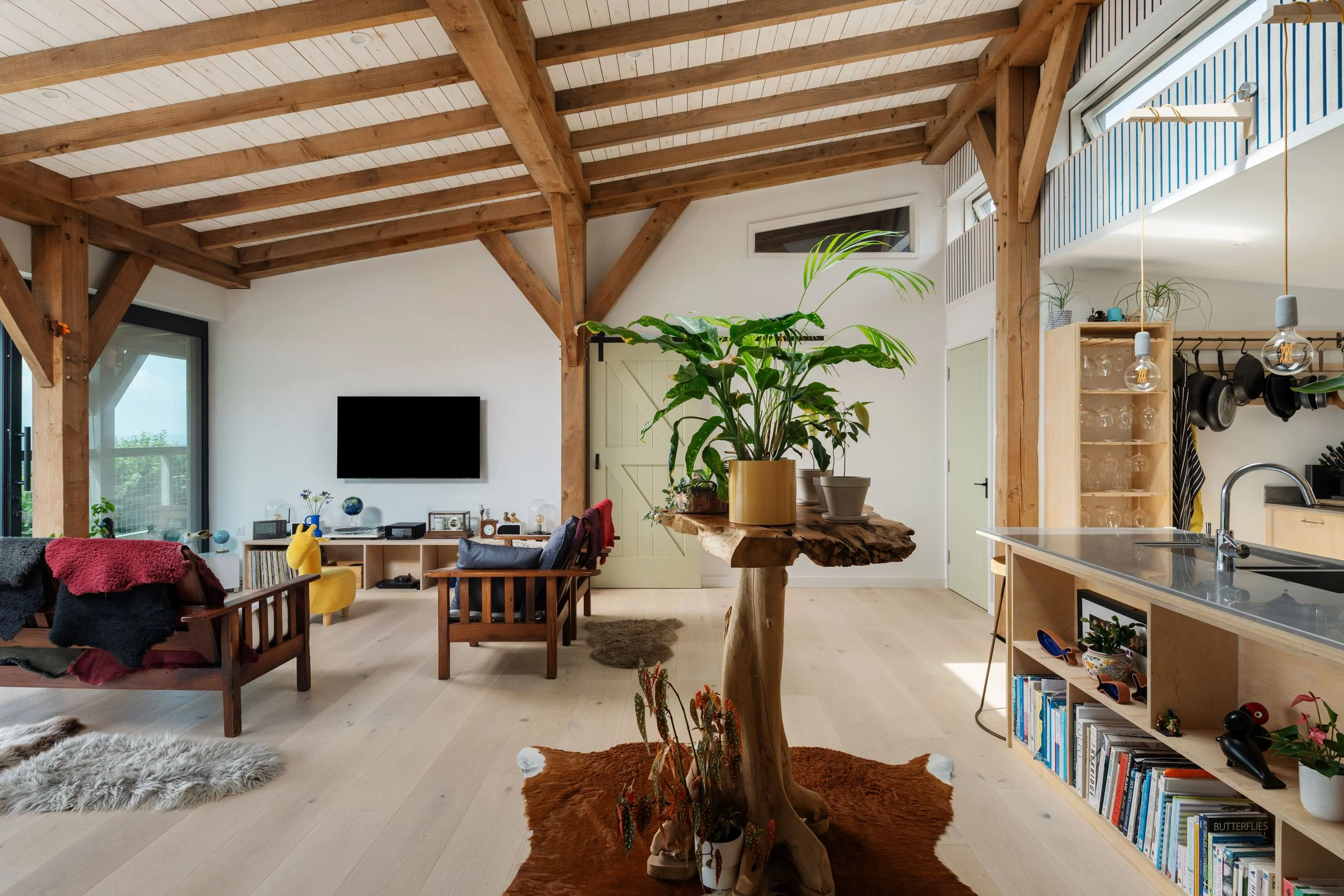 Living room with wooden ceiling beams, large window, and houseplants, featuring a sitting area with wooden chairs, a small table, and a bookshelf.