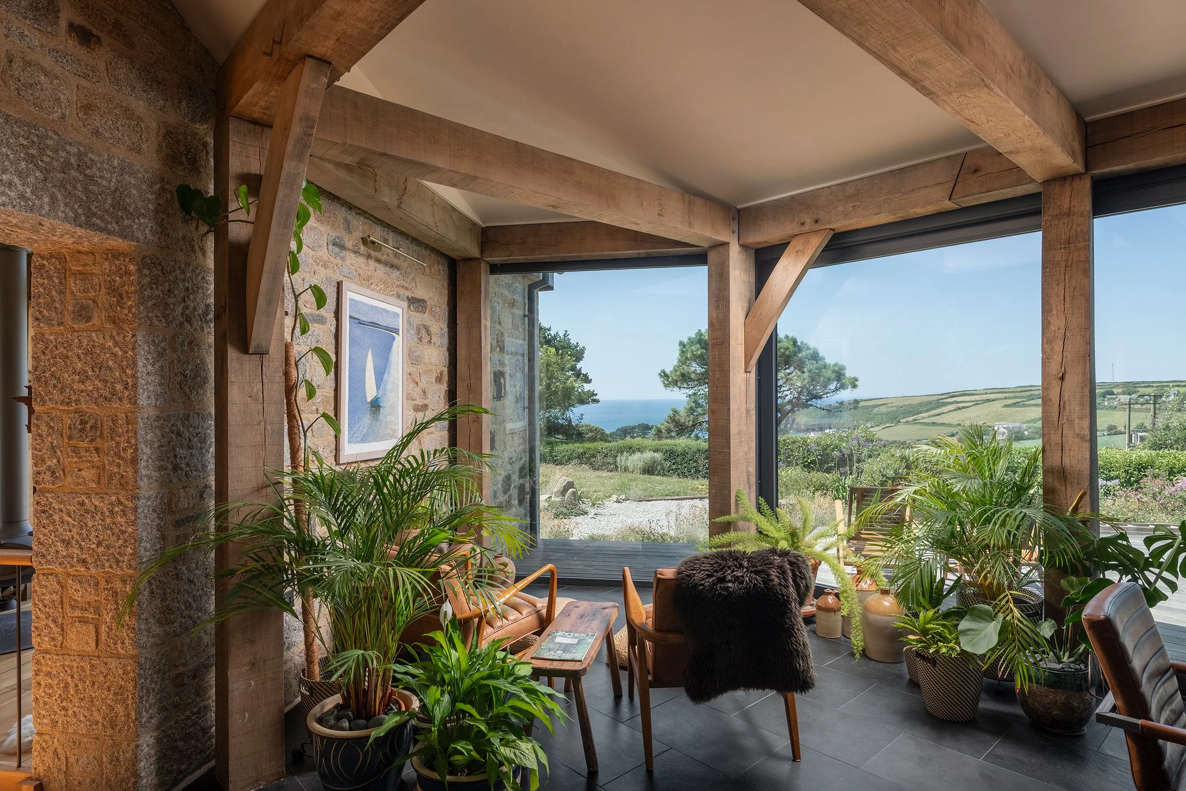 Roderick James Architects LLP — Interior of a room with large glass windows showing an outdoor landscape with trees and hills, decorated with wooden beams, stone walls, and numerous