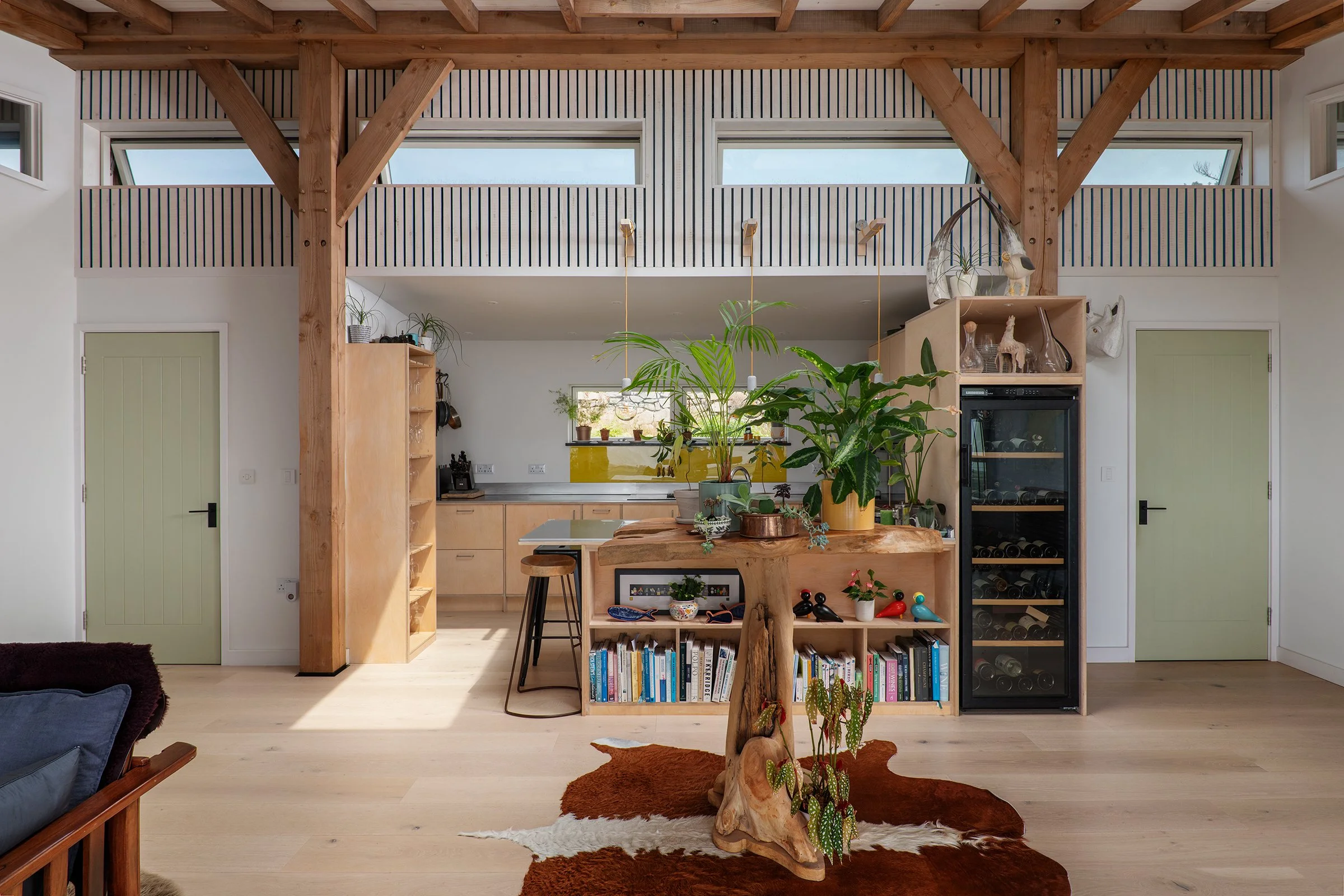 Modern open-concept kitchen and living area with wooden beams, a bookshelf, indoor plants, and large windows.
