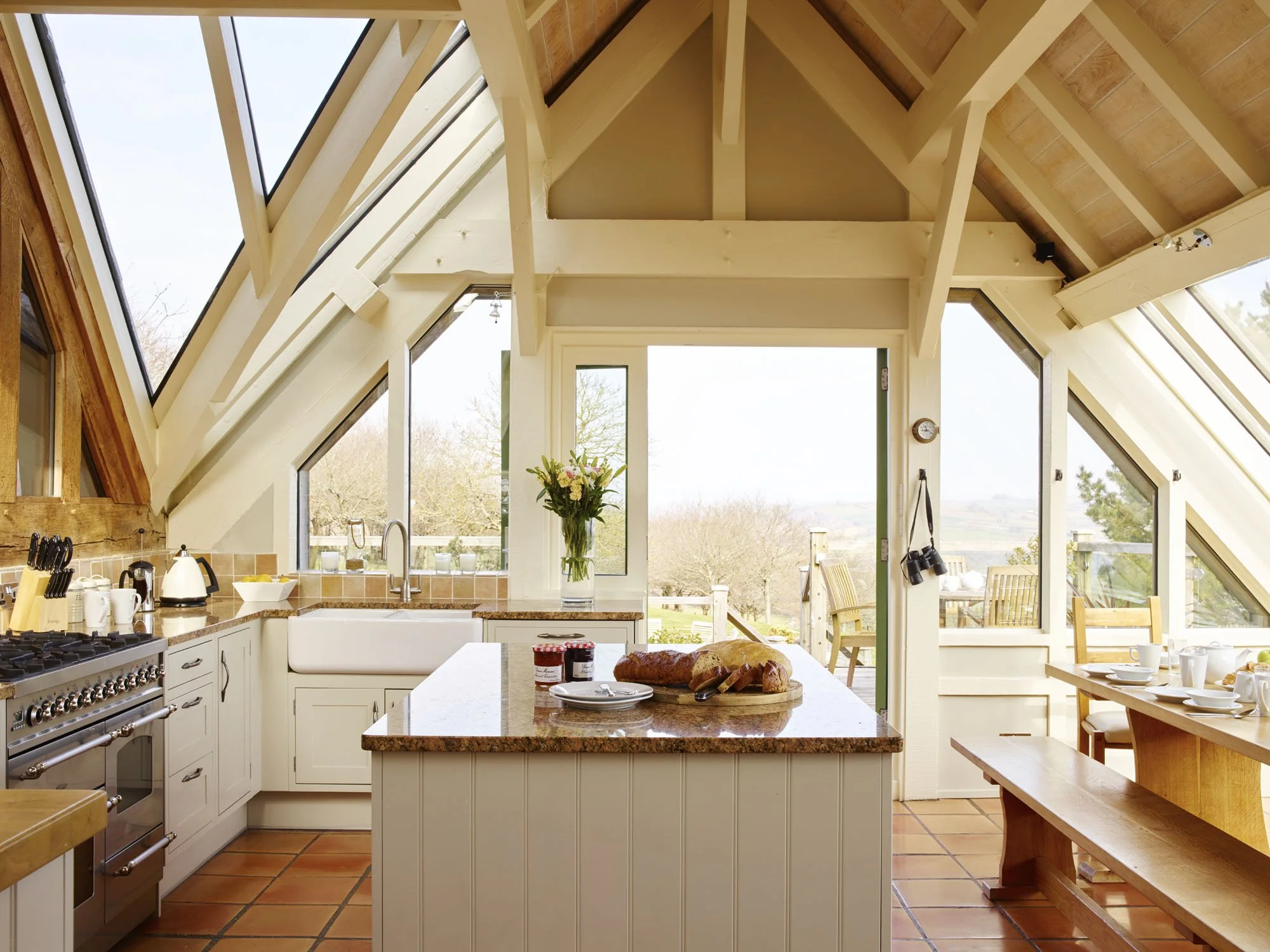 A bright, airy kitchen with a skylight, large windows, and a door leading to an outdoor patio. There is a kitchen island with a loaf of bread, jam, and plates. The countertops have a vase with flowers and kitchen appliances, including a stove, kettle