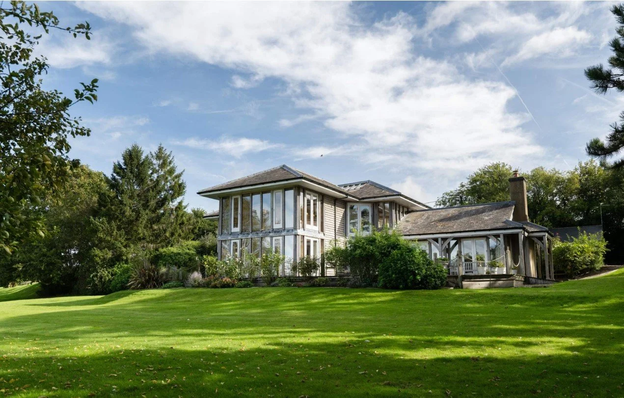 A large house with multiple glass windows on a grassy lawn with trees and a partly cloudy sky.