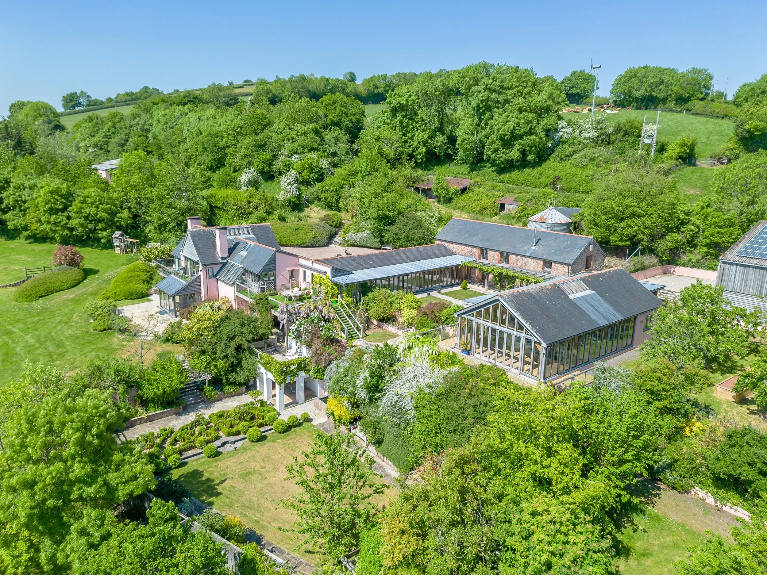 Aerial view of a lush green hillside with houses, gardens, and trees, under a clear blue sky, including wind turbines on the hilltop.