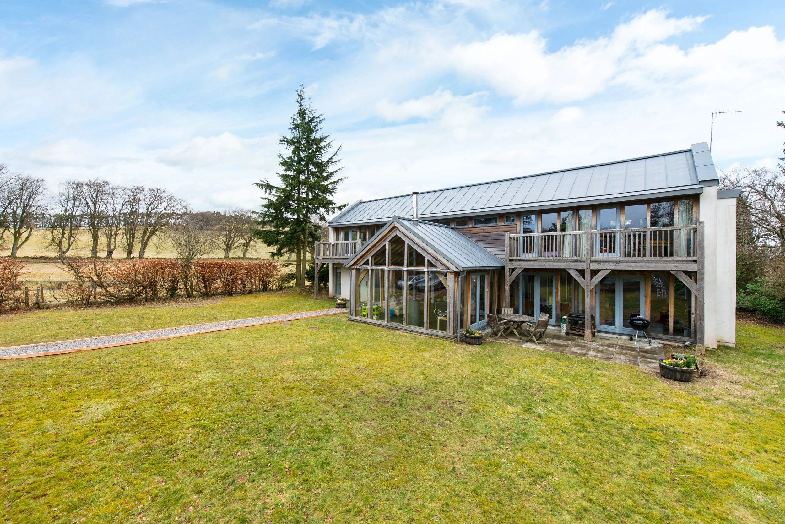 A modern two-story house with a glass conservatory in a large grassy yard, surrounded by a hedge and trees, under a partly cloudy sky.