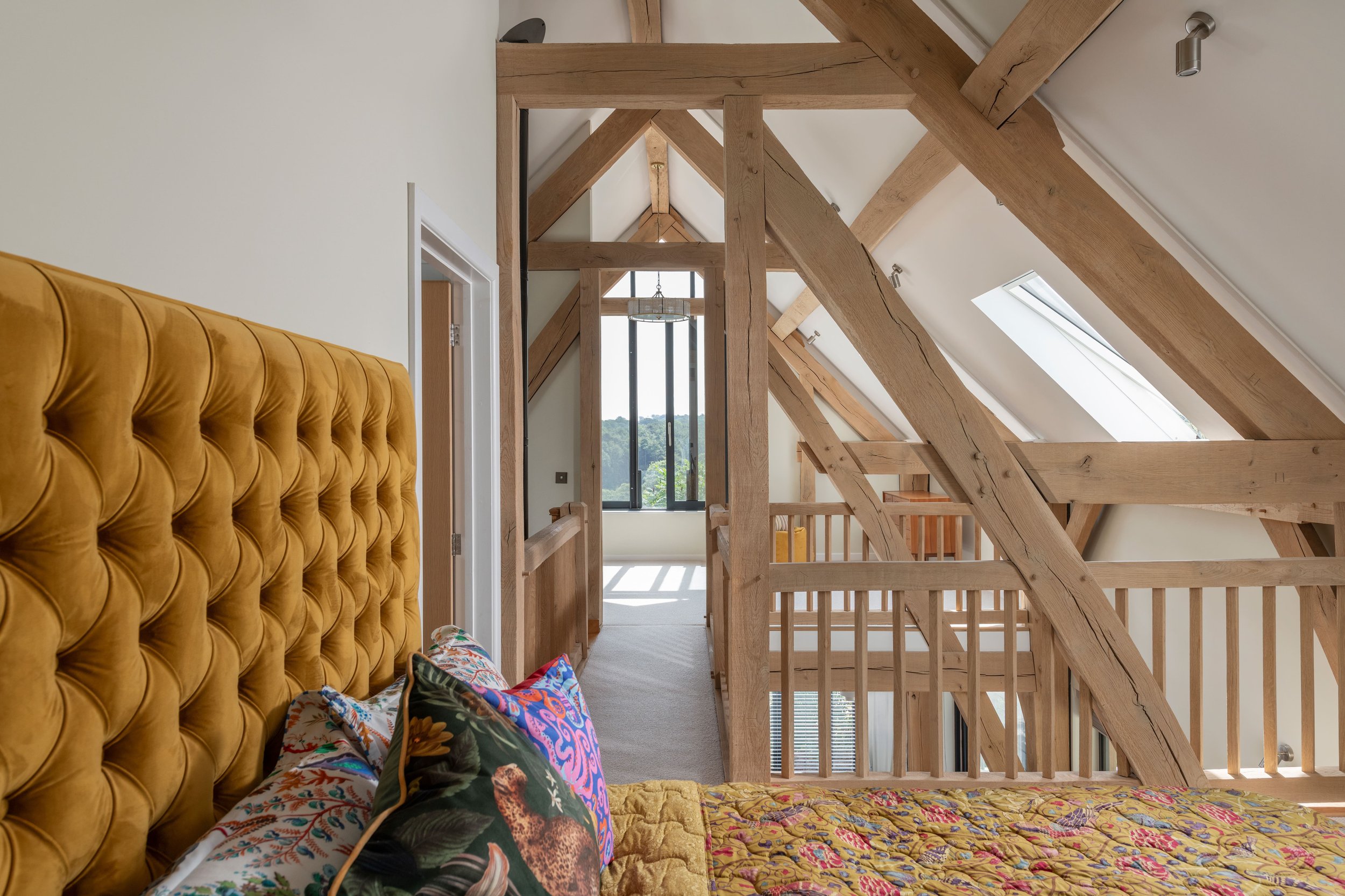 Interior of a house with exposed wooden beams, skylight windows, a mustard-colored tufted headboard, colorful patterned bedding, and decorative pillows. Wooden stairs and railing are visible, with a view of trees outside through glass doors.
