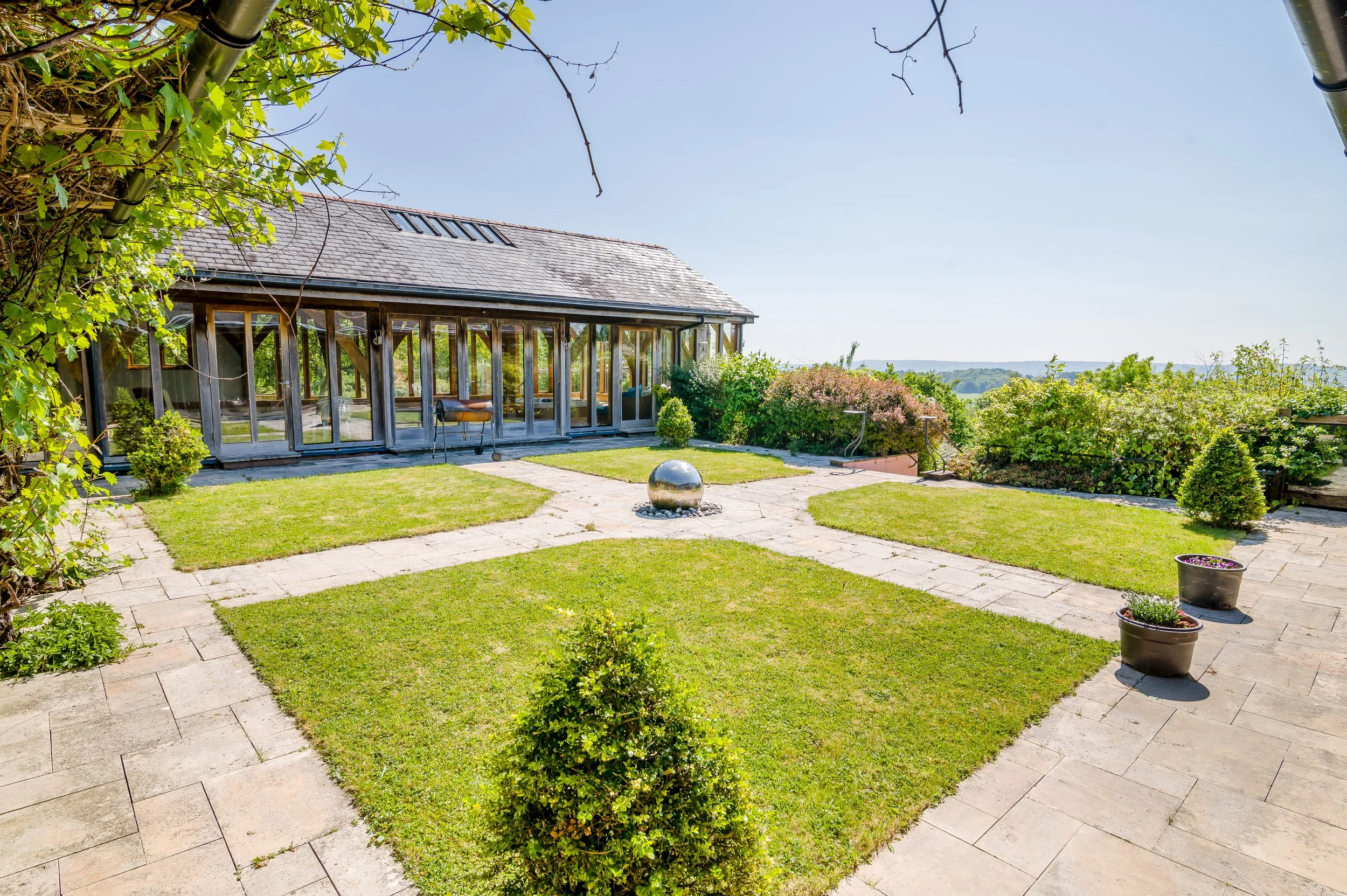 A sunny backyard with a well-maintained lawn and paved pathways, surrounded by various bushes and plants. There is a glass-enclosed porch attached to a house with a shingled roof and a row of skylights. A large reflective metal sphere sculpture is in