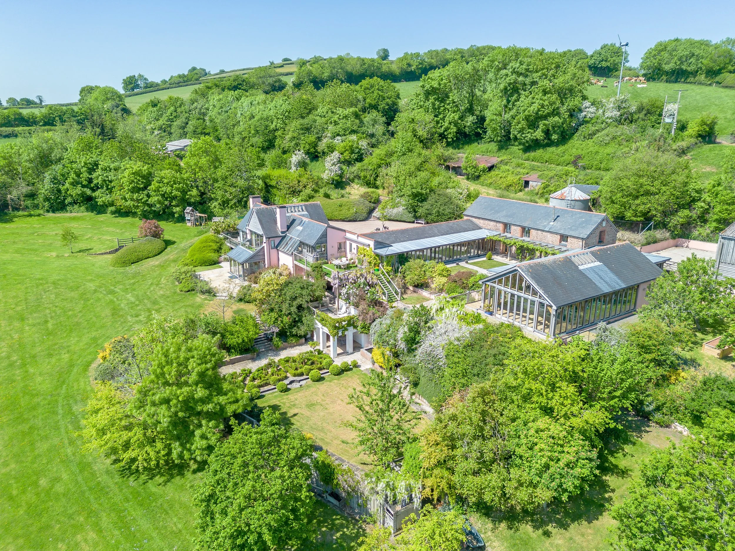 Aerial view of a large house surrounded by lush green trees, rolling hills, and open grassy areas on a bright sunny day.