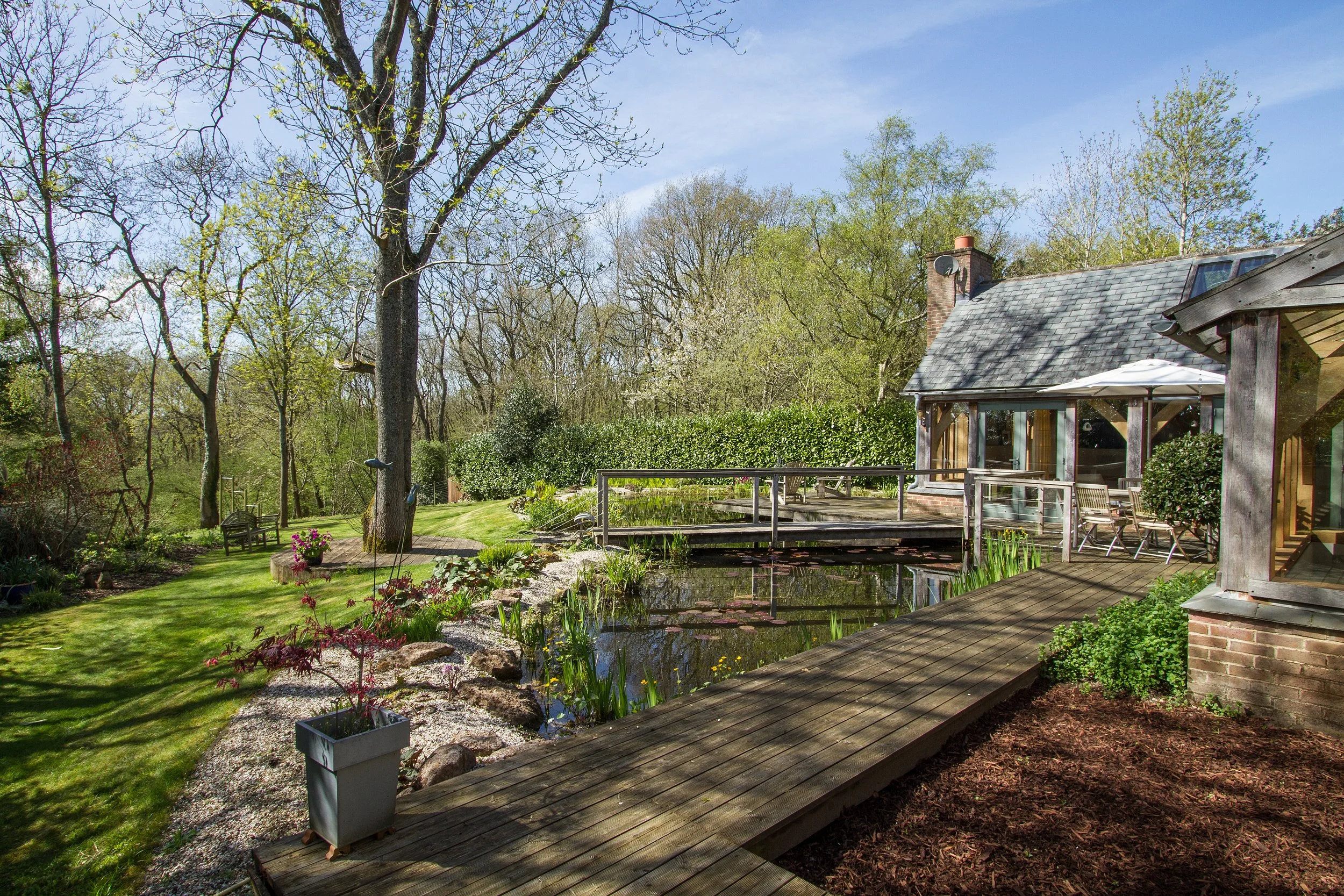 A backyard garden with a pond, wooden deck, patio furniture, and a house with a chimney and solar panels on the roof, surrounded by trees and greenery.