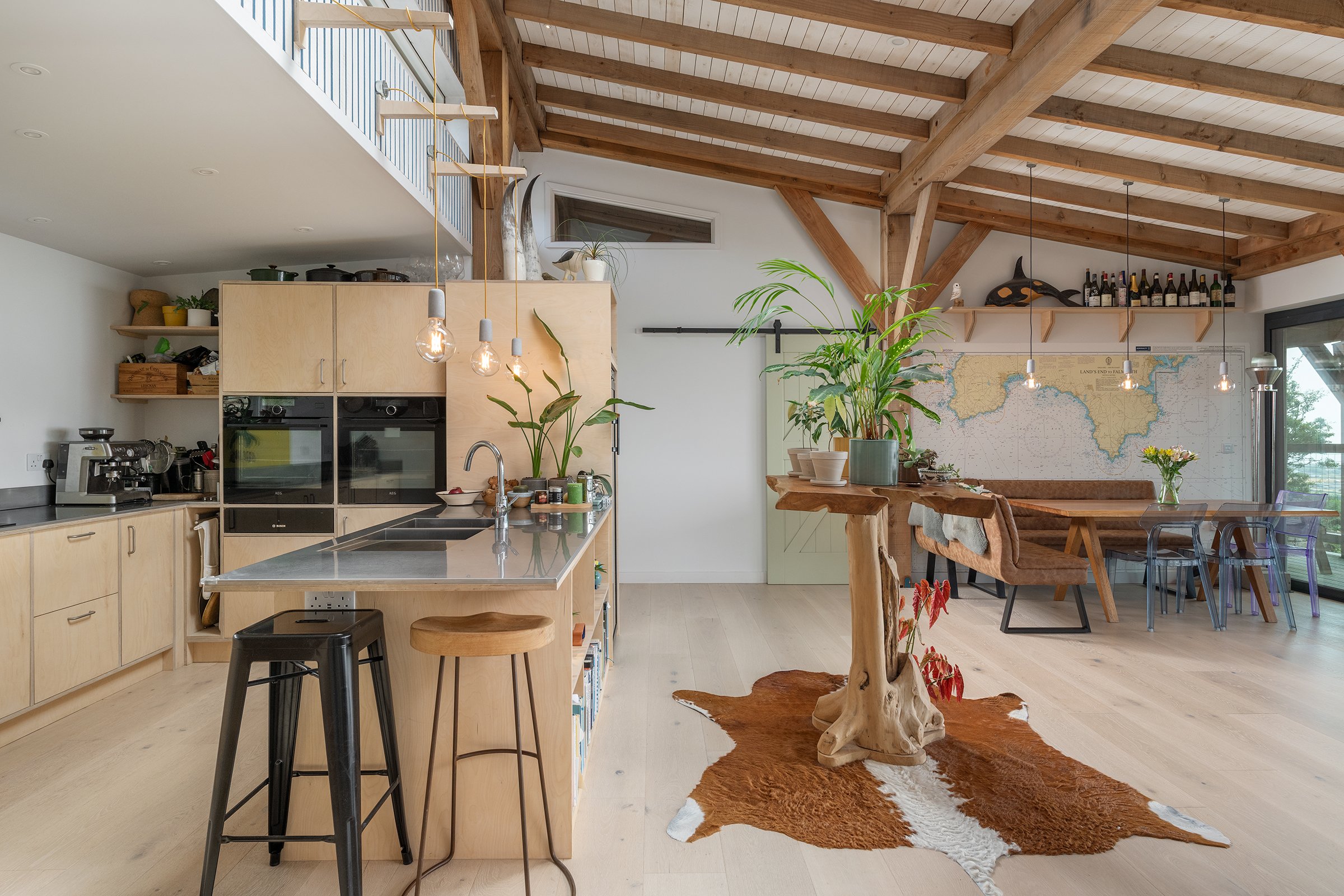 Open-concept kitchen and dining area with wooden beams and vaulted ceiling. Kitchen has light wood cabinetry, black appliances, and a kitchen island with a sink, decorated with plants and small items. Hanging pendant lights above the island. Dining a