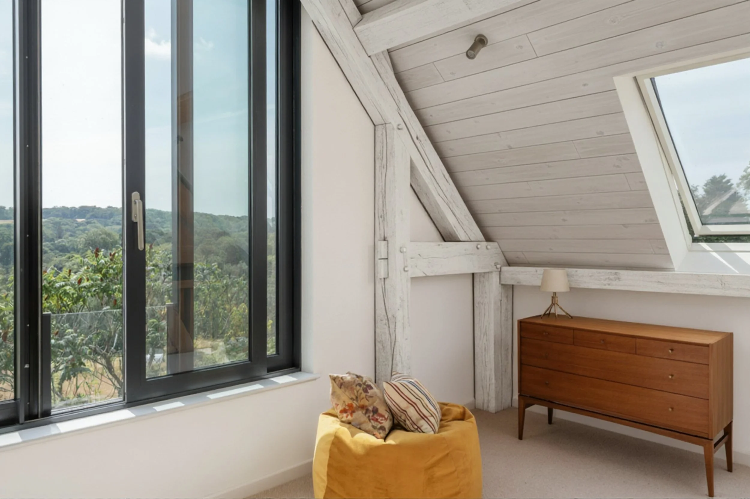 A room with a large window showing greenery outside, a wooden dresser, a yellow armchair with pillows, and a sloped ceiling with a skylight.