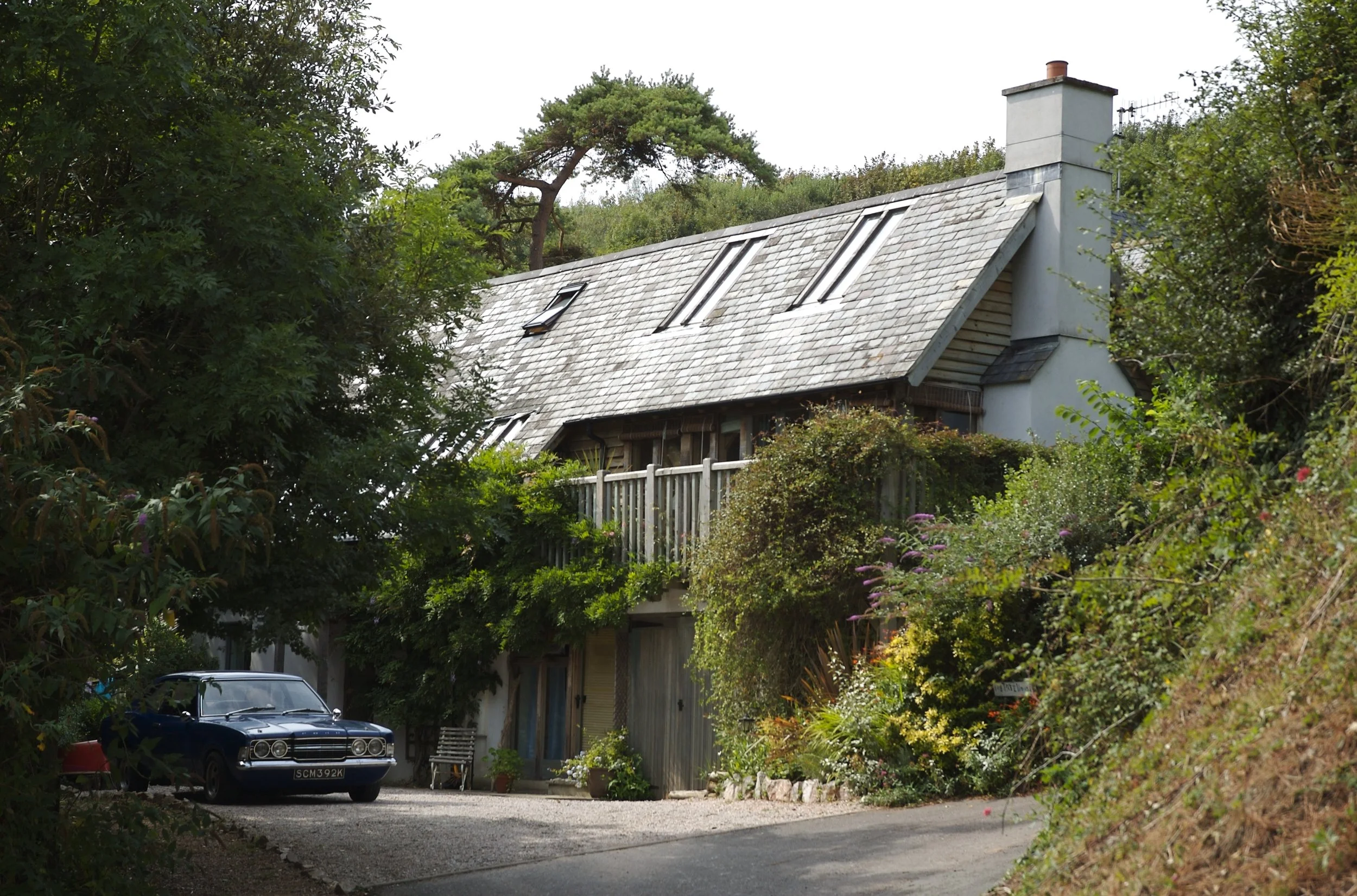 A white, two-story house with a steeply sloped roof, multiple skylights, and a chimney. The house is surrounded by dense green foliage and trees. A classic black car is parked in front, along with some garden décor and potted plants.