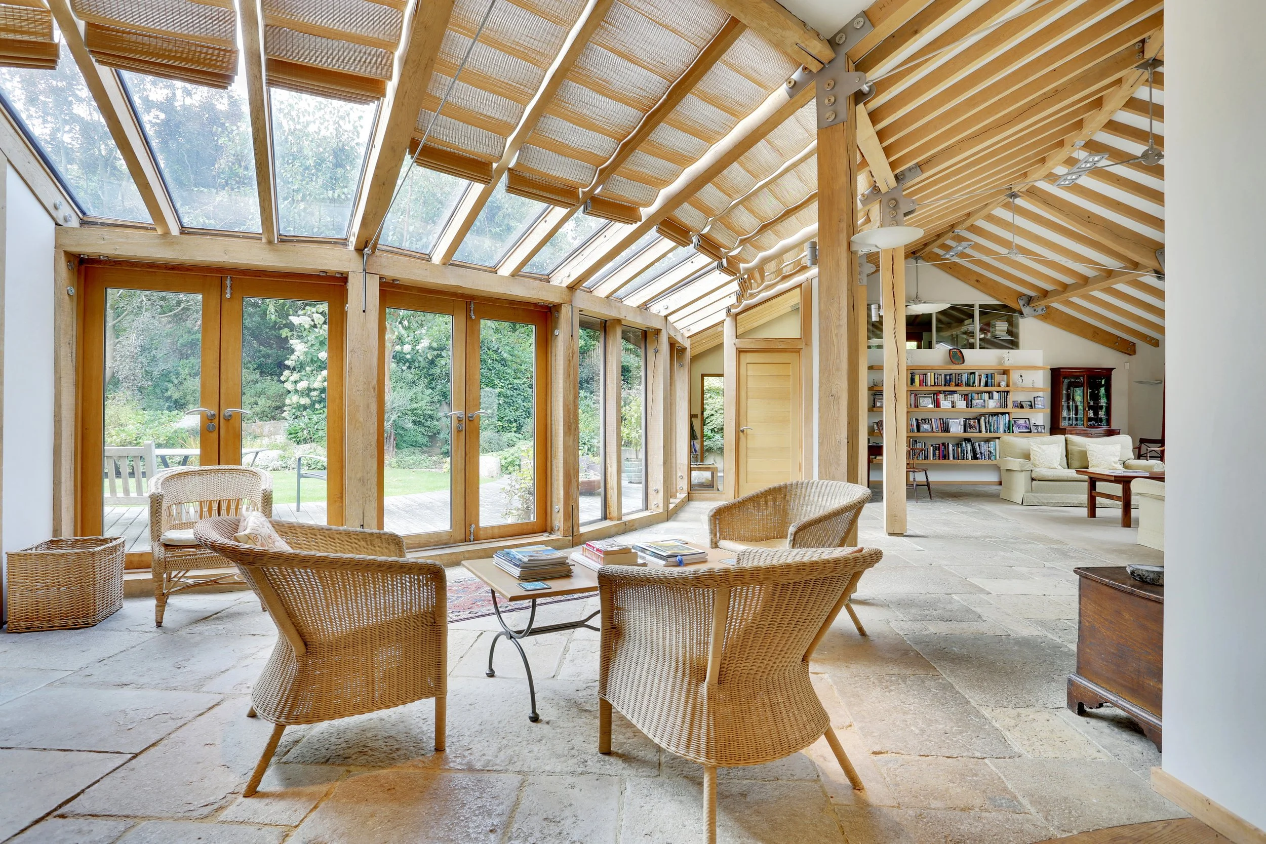Sunroom with exposed timber beams, glass roof, wicker chairs, coffee table, garden views, and adjoining living room with bookshelves