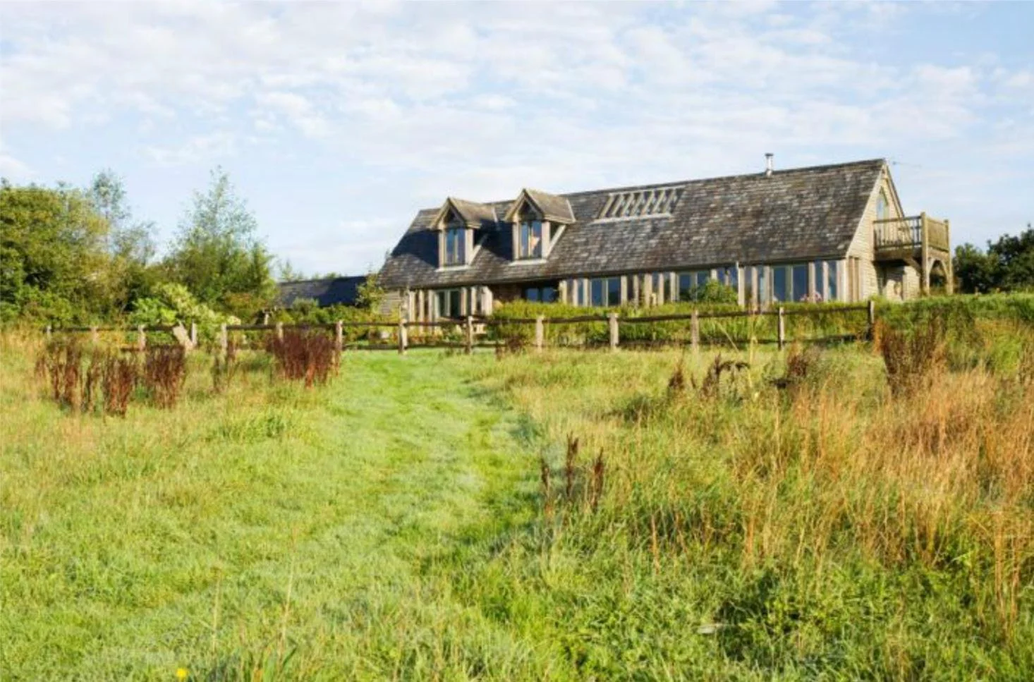 A rustic house with a timber exterior and a steep shingle roof sits on a grassy hill, surrounded by a wooden fence and overgrown grass, with trees and a blue sky in the background.
