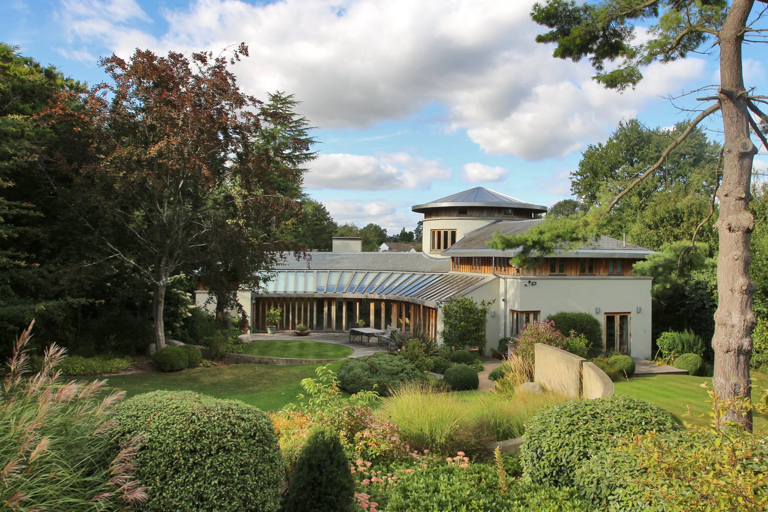 Modern house with glass-roofed extensions surrounded by lush landscaped gardens under a partly cloudy sky