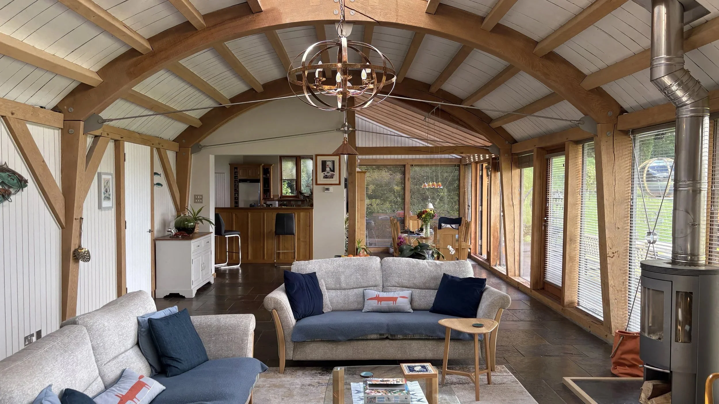 Living room with exposed wooden beams, large windows with blinds, gray sofa, decorative pillows, a coffee table with magazines, and a modern chandelier hanging from the ceiling.
