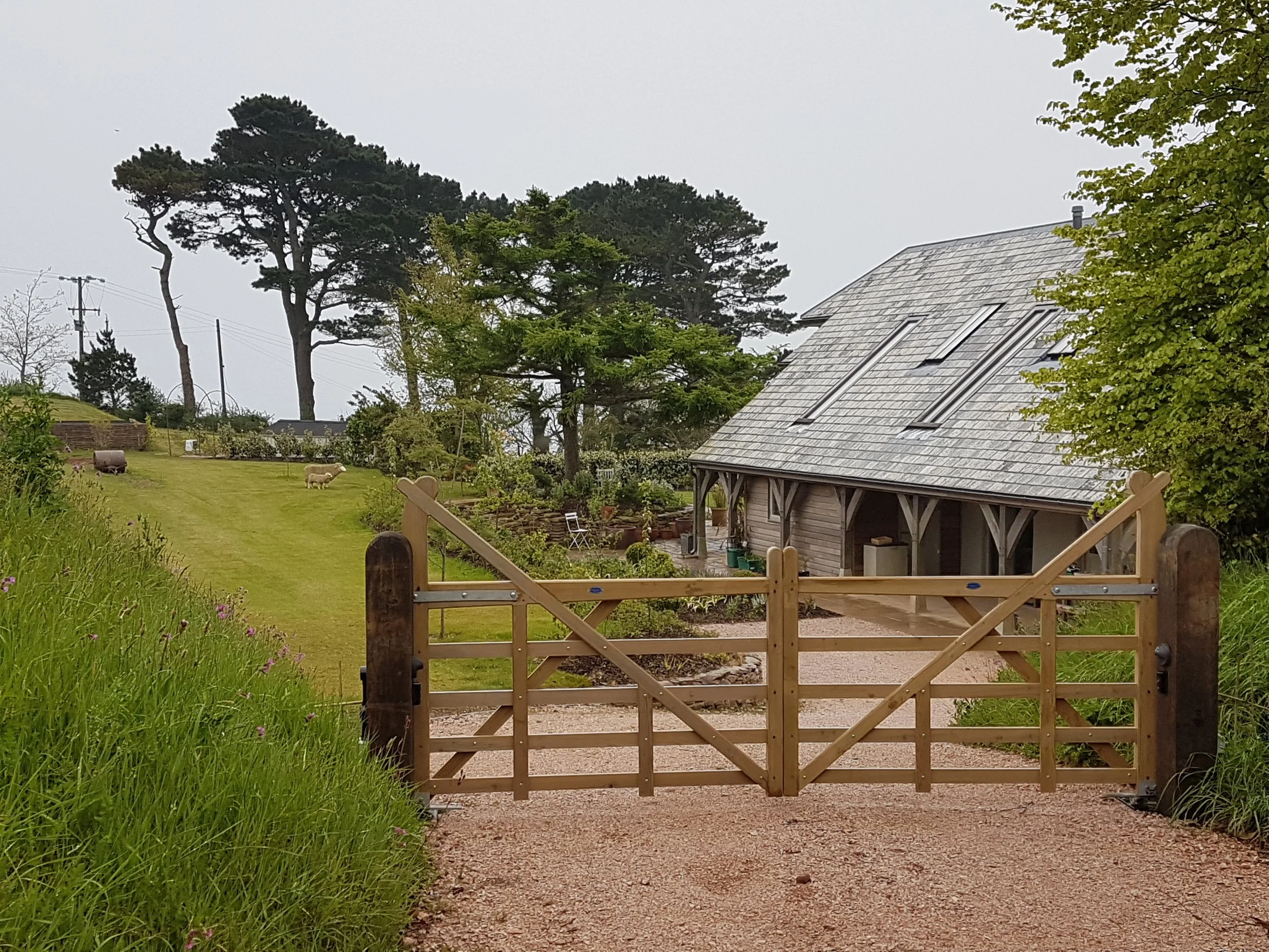 A wooden gate leading to a garden with a rustic house, grazing sheep, and tall trees in the background on a cloudy day.