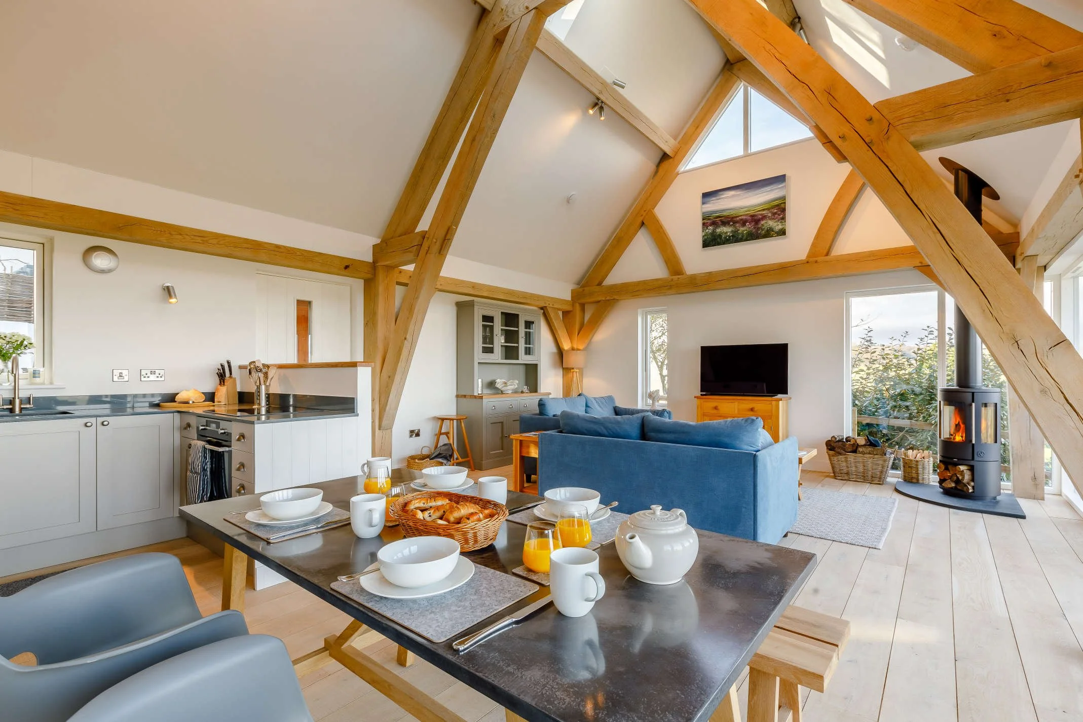 Open-concept living and dining area with exposed wooden beams, a modern fireplace, large windows, and a kitchen with white cabinets and black countertop.