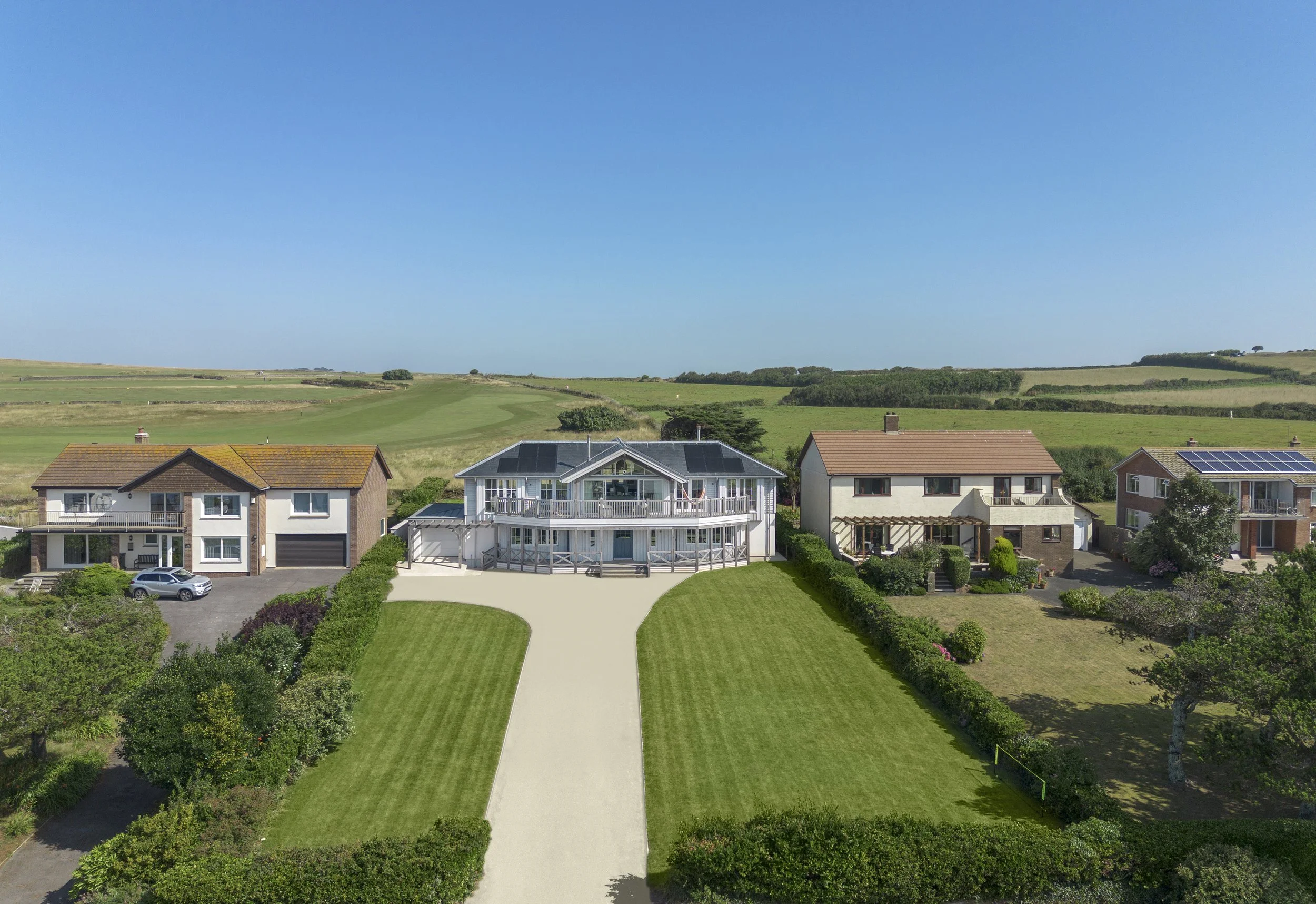 Aerial view of a row of houses with green lawns and hedges, set against a rural landscape with open fields and a clear blue sky.