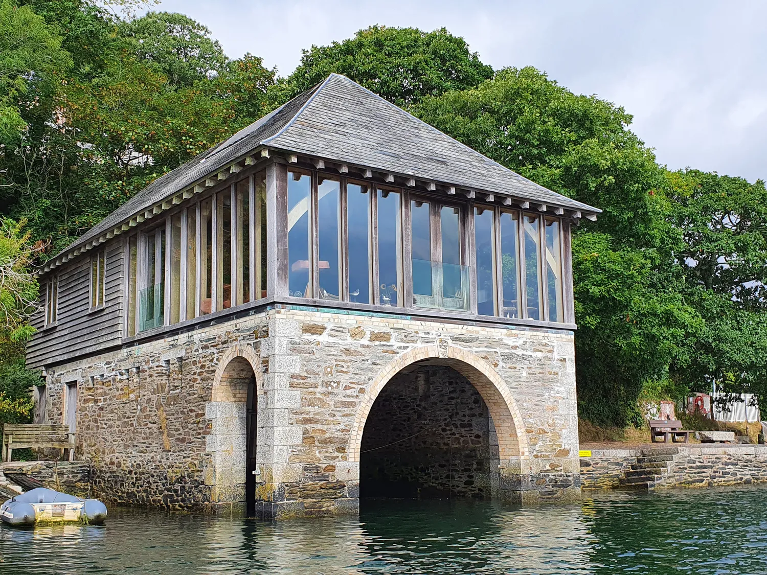 A two-story building with a stone archway on the water, featuring large windows on the upper level, surrounded by green trees.