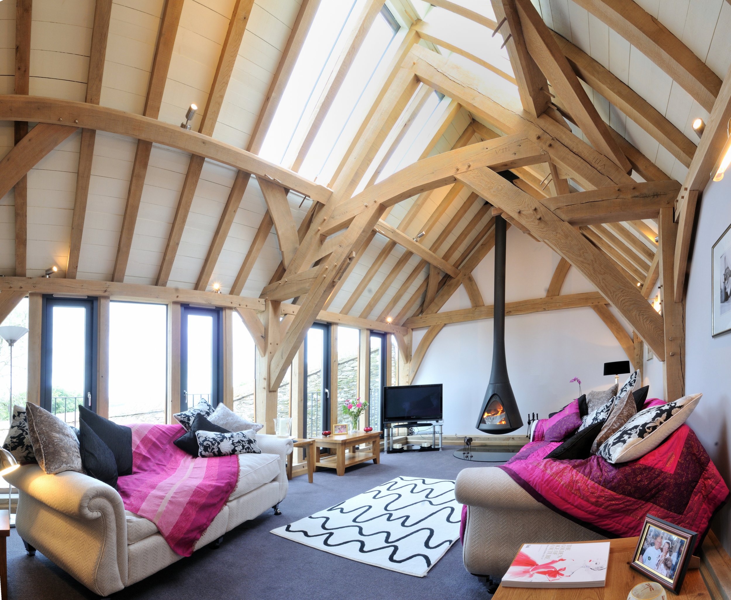 Living room with high ceiling, exposed wooden beams, large windows, fireplace, and seating area with colorful cushions and a black and white rug.