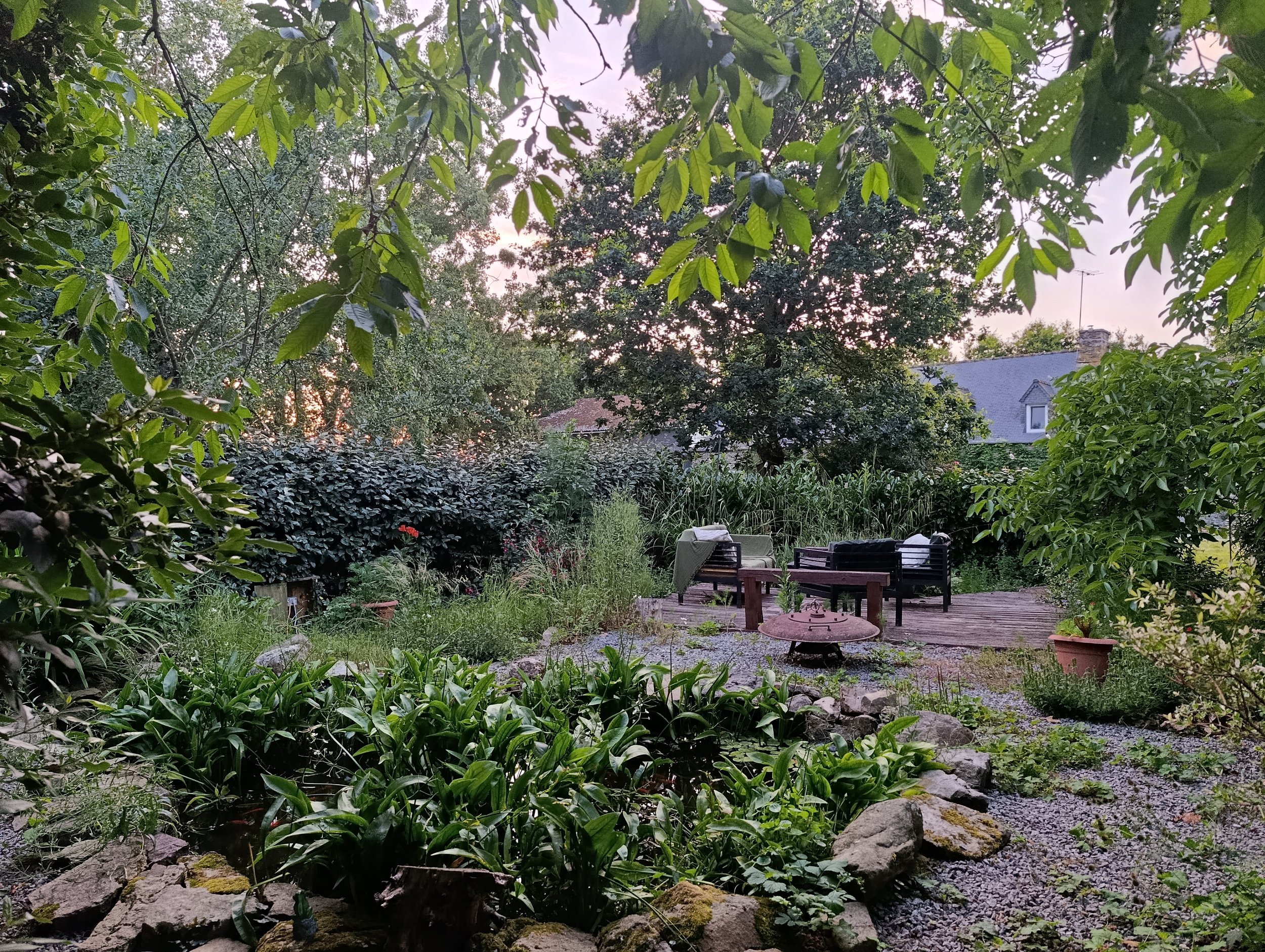 een tuinhoekje omgeven door fruitbomen met een vijver, een zitplek en een vuurkorf in bretagene met een ondergaande zon- chambres dhotes