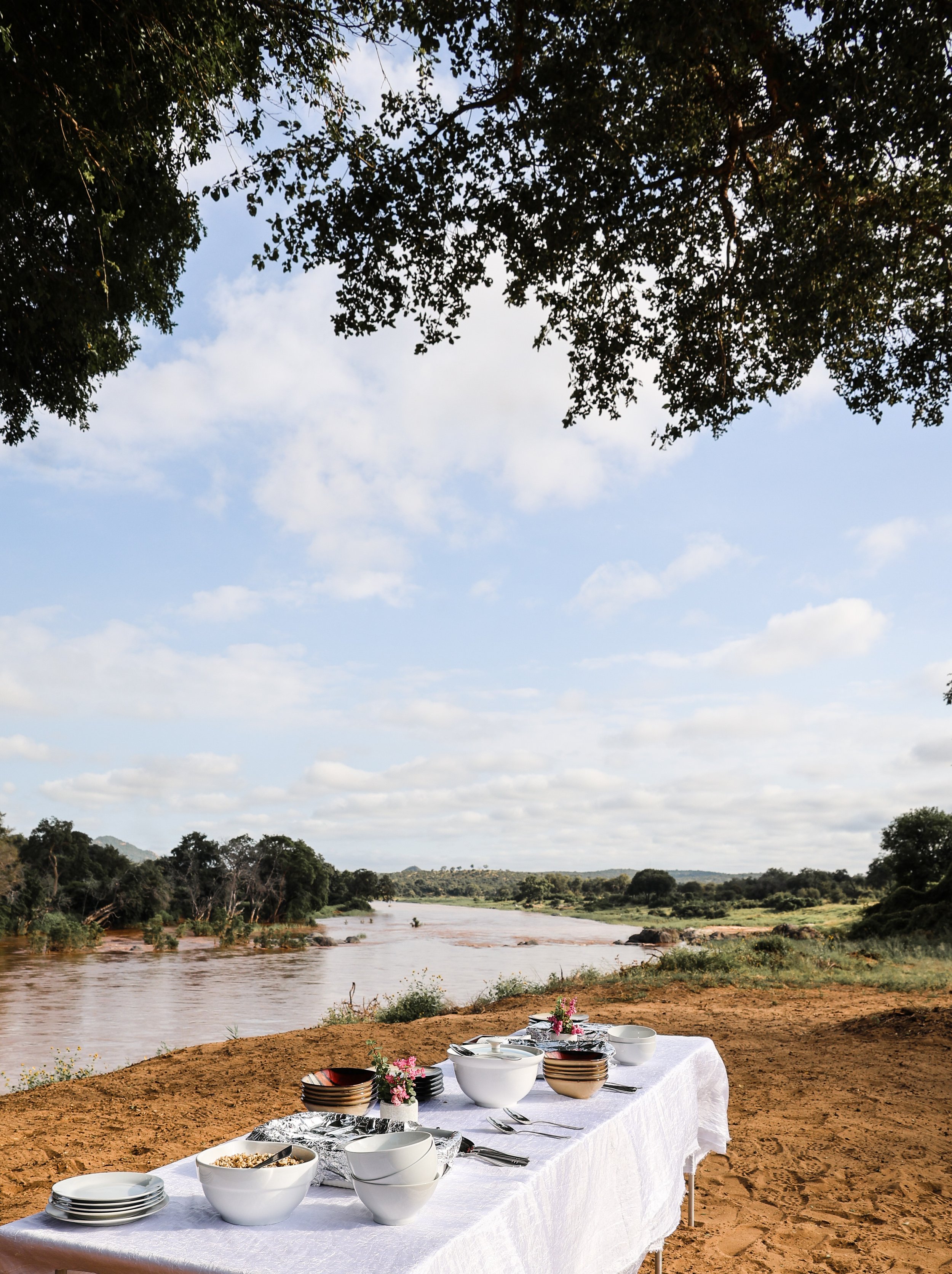 Outdoor dining table set with bowls, plates, and utensils on a white tablecloth, near a river with trees and blue sky in the background.