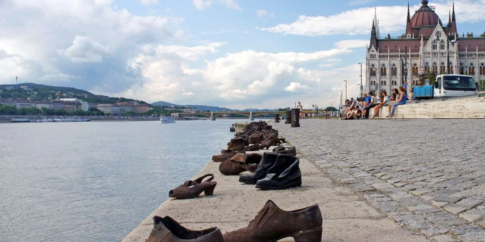 Shoes and boots lined up along a riverbank with city buildings, bridge, and people sitting on the edge near a historic building.