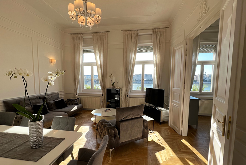 Bright living room with three large windows, beige curtains, a gray sofa, a brown velvet armchair, a television, and a dining table with orchids in a pot.