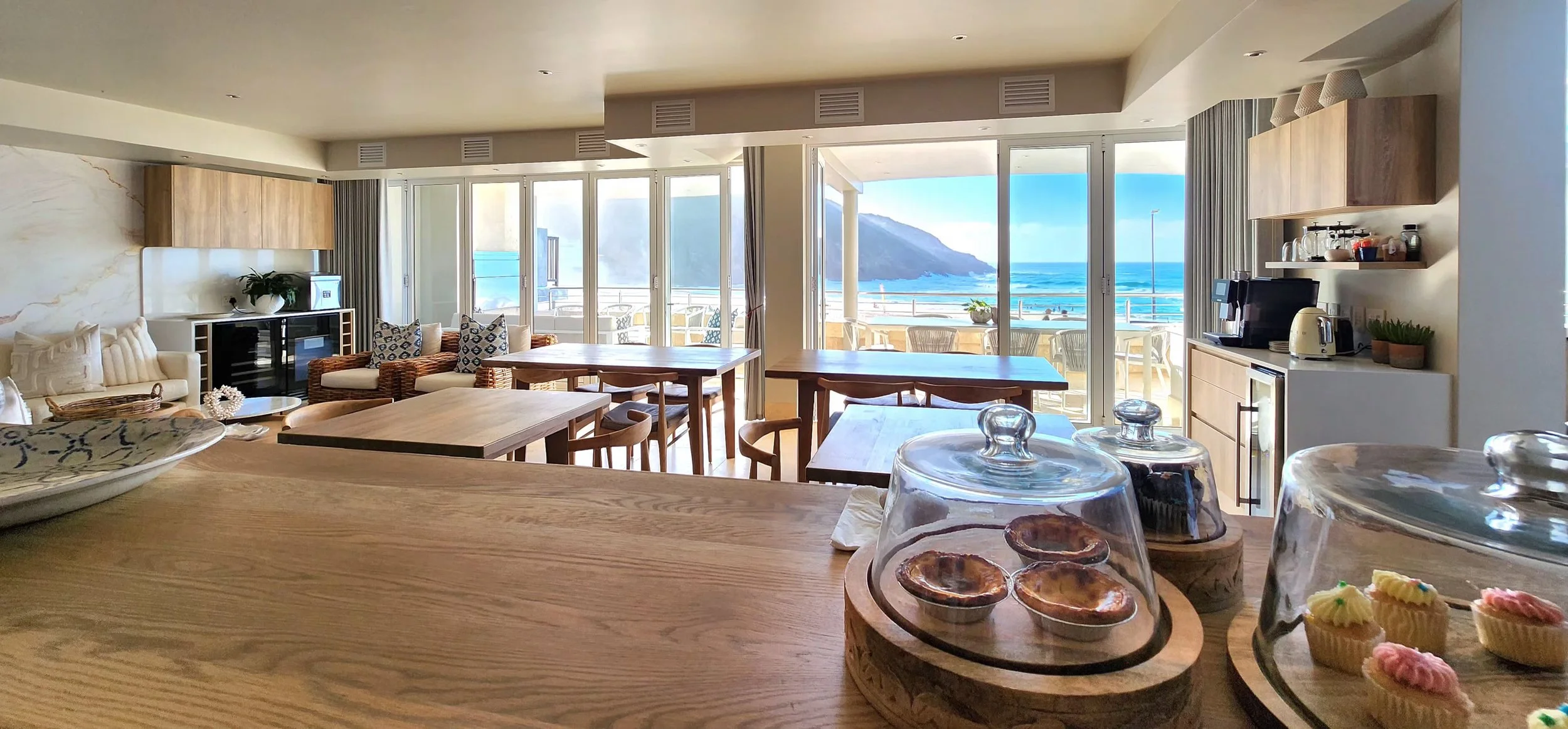 Living room with sea view, outdoor patio with chairs, kitchen area with coffee maker, toaster, and cupcakes under glass domes, wooden tables and chairs, and seating area with cushions.