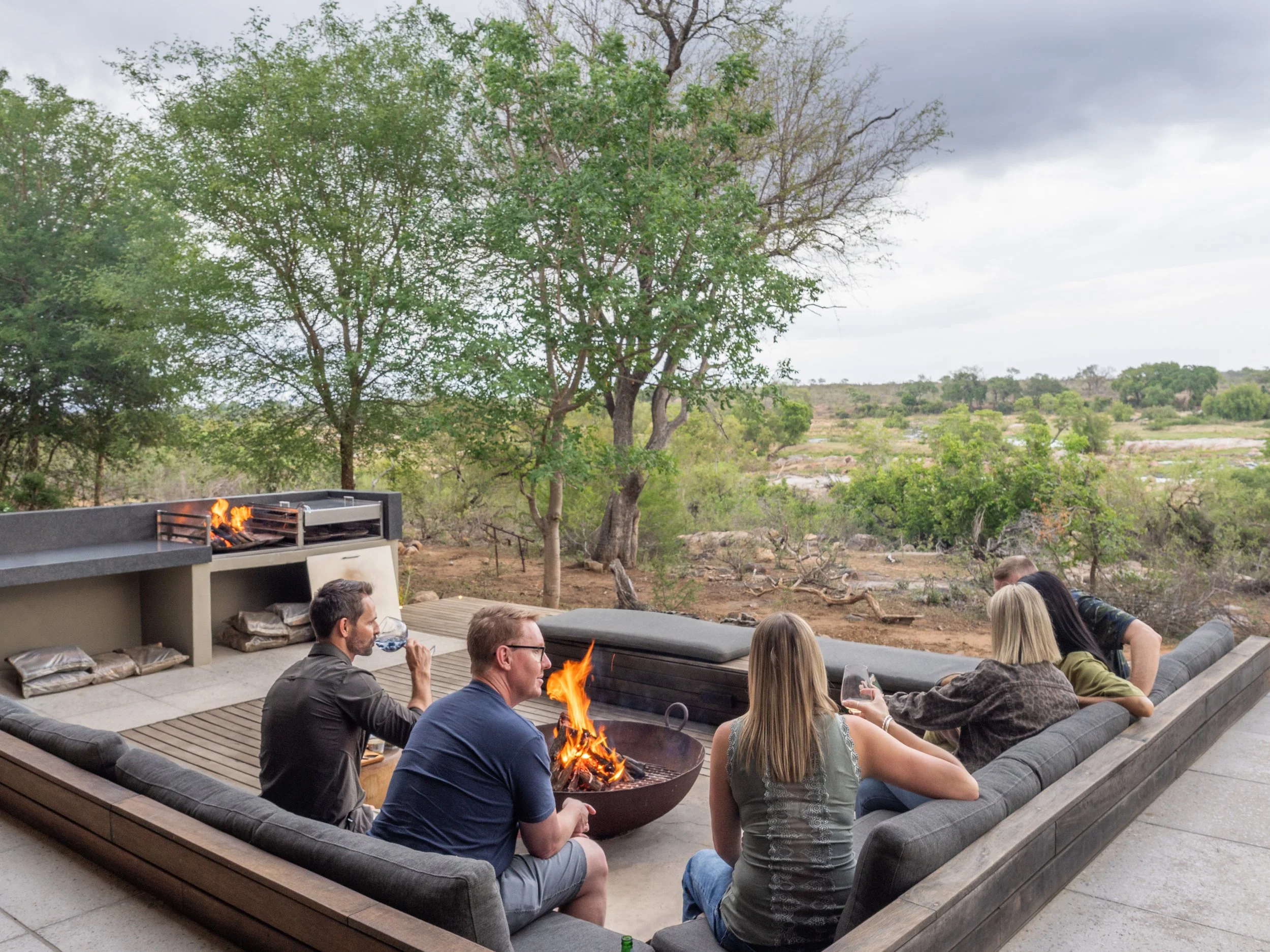 Group of six friends sitting around a fire pit on outdoor patio, enjoying drinks and conversation, with a backyard landscape of trees and open land, and a built-in gas grill in the background