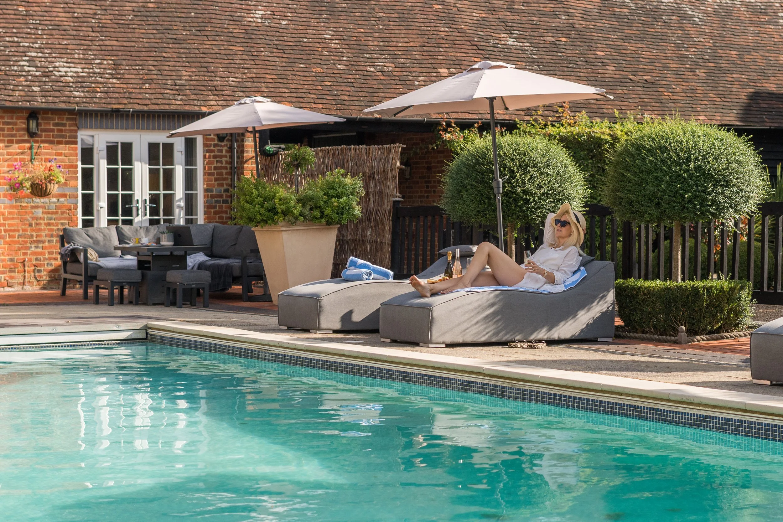 A woman with blonde hair and sunglasses relaxing on a lounge chair beside a swimming pool, with a bottle of wine and a glass. Two large umbrellas shade the area, with a brick house and green bushes in the background.