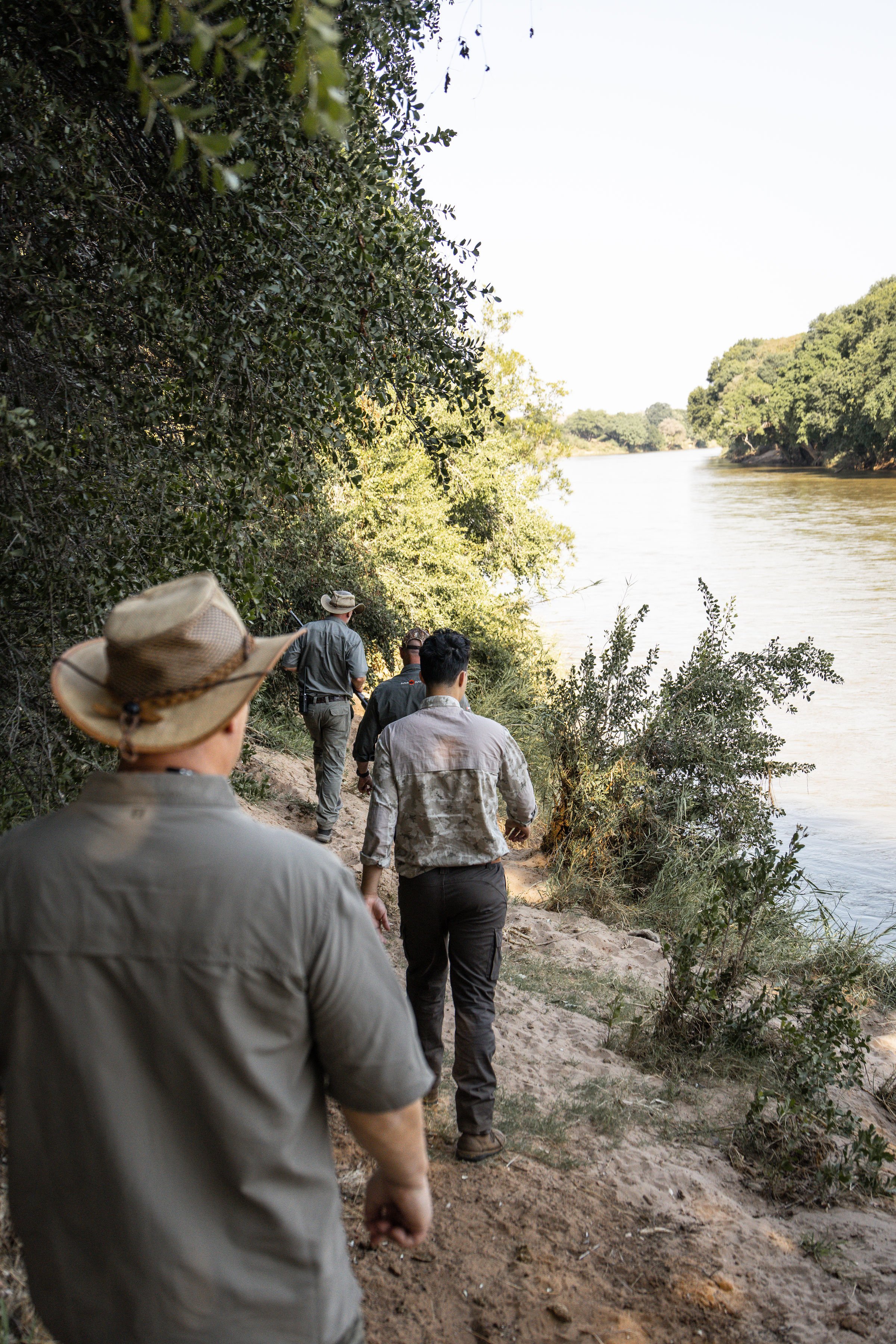 A group of people walking along a dirt path beside a river, surrounded by trees and foliage.