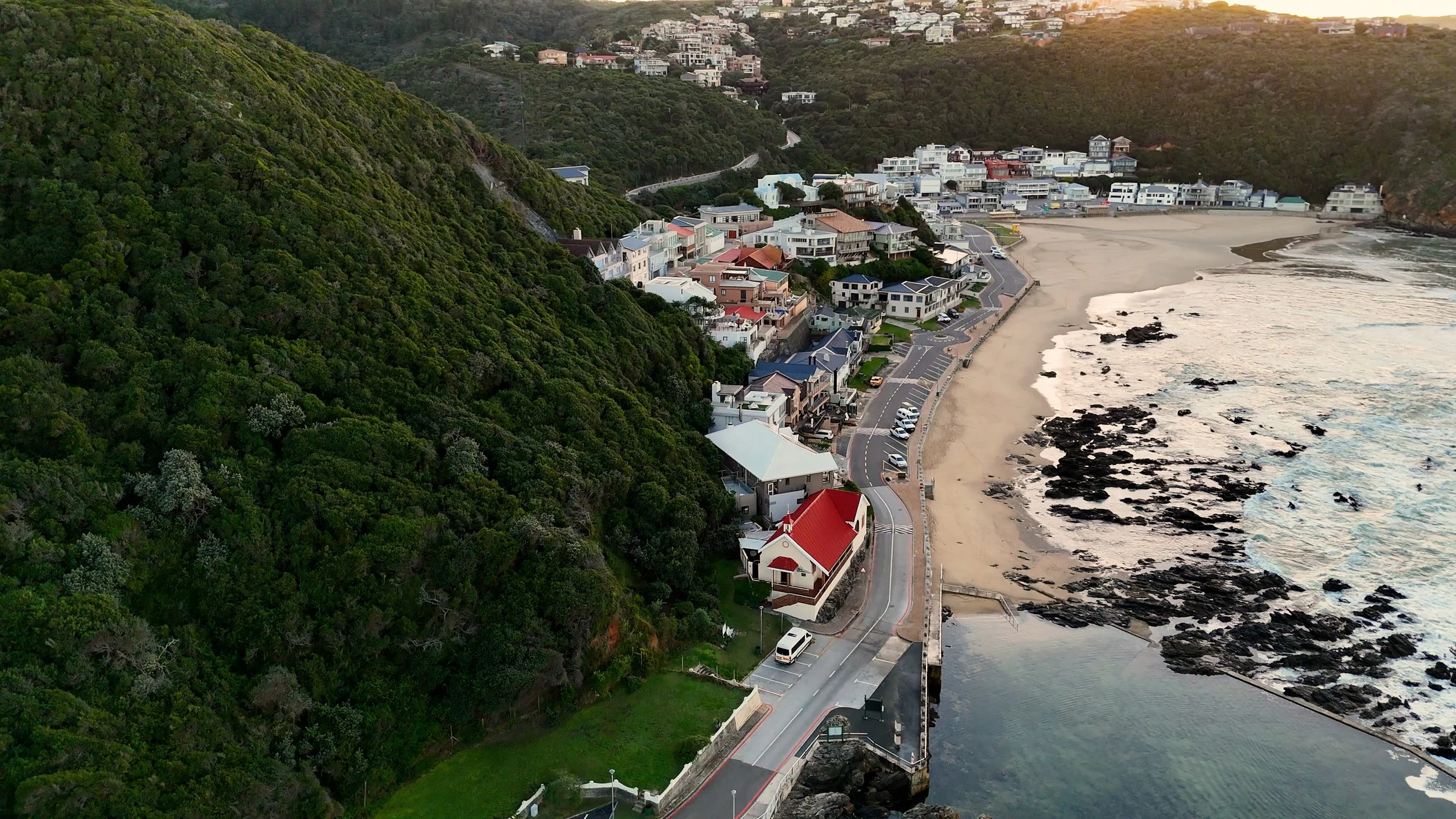 Aerial view of a coastal town with colorful houses on a hillside next to a sandy beach and rocky shoreline during sunset.