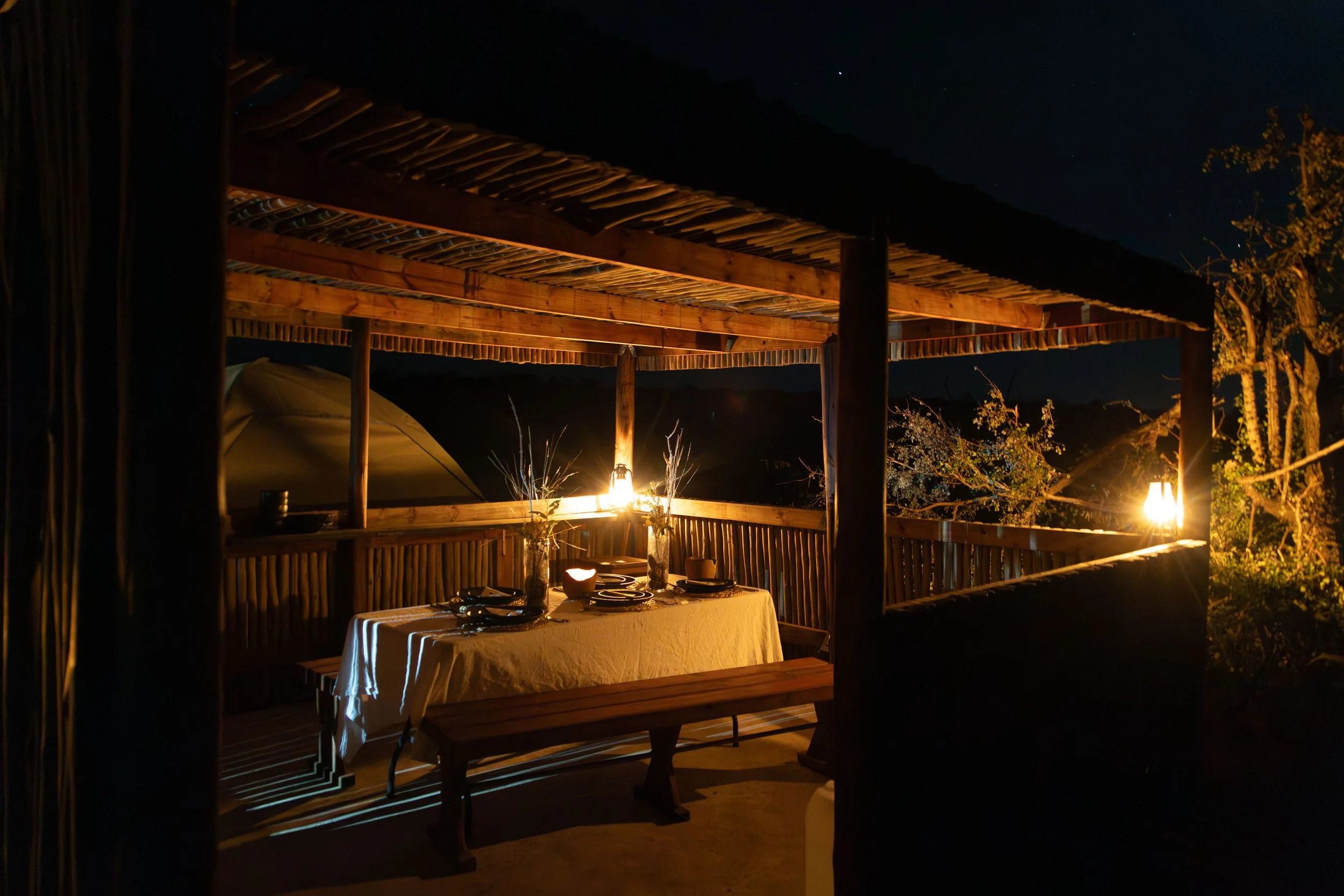 An outdoor wooden dining area at night with a table set for a meal, illuminated by lanterns, surrounded by trees and mountains in the background.