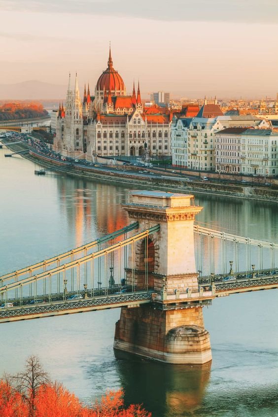 A cityscape featuring the Hungarian Parliament building and the Chain Bridge over the Danube River in Budapest, Hungary, during sunset.