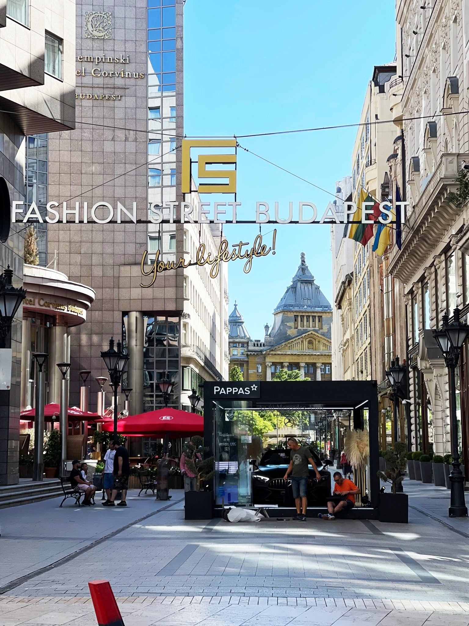 Street view in Budapest with large signs reading 'Fashion Street Budapest' and 'Your lifestyle!', a black booth labeled 'Pappas', pedestrians, outdoor seating with red umbrellas, historic and modern buildings, flags, and a bright blue sky.