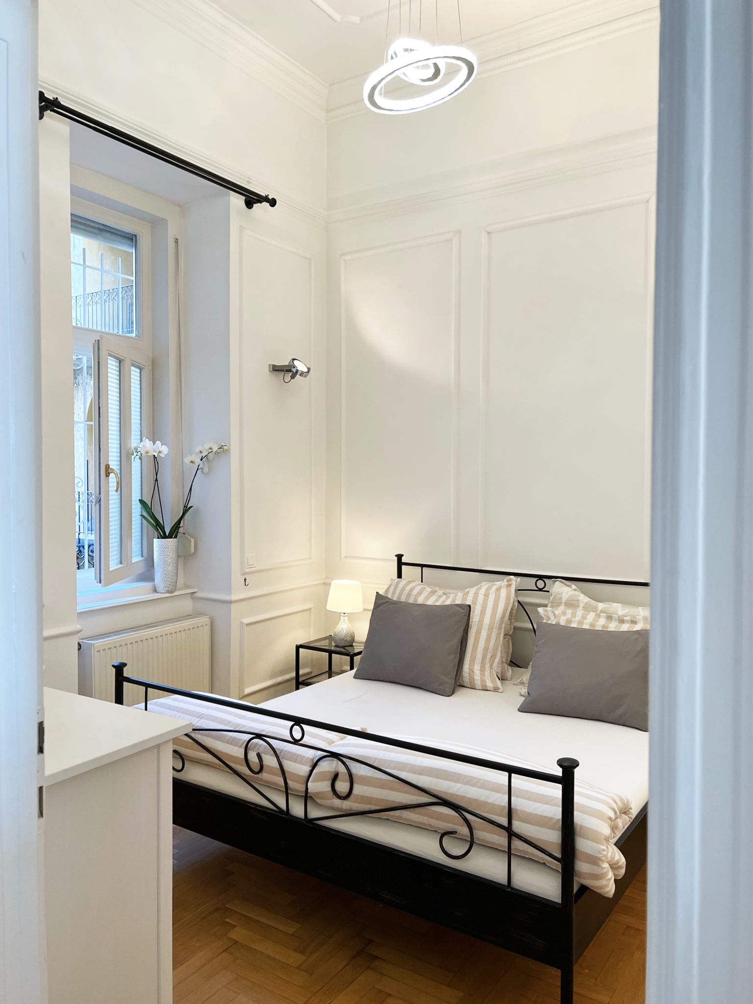 A bedroom with white walls, black metal bed frame, striped bedding, gray and striped pillows, a small white nightstand with a lamp, a window with open shutters, a vases with white orchids, and modern ceiling lighting.