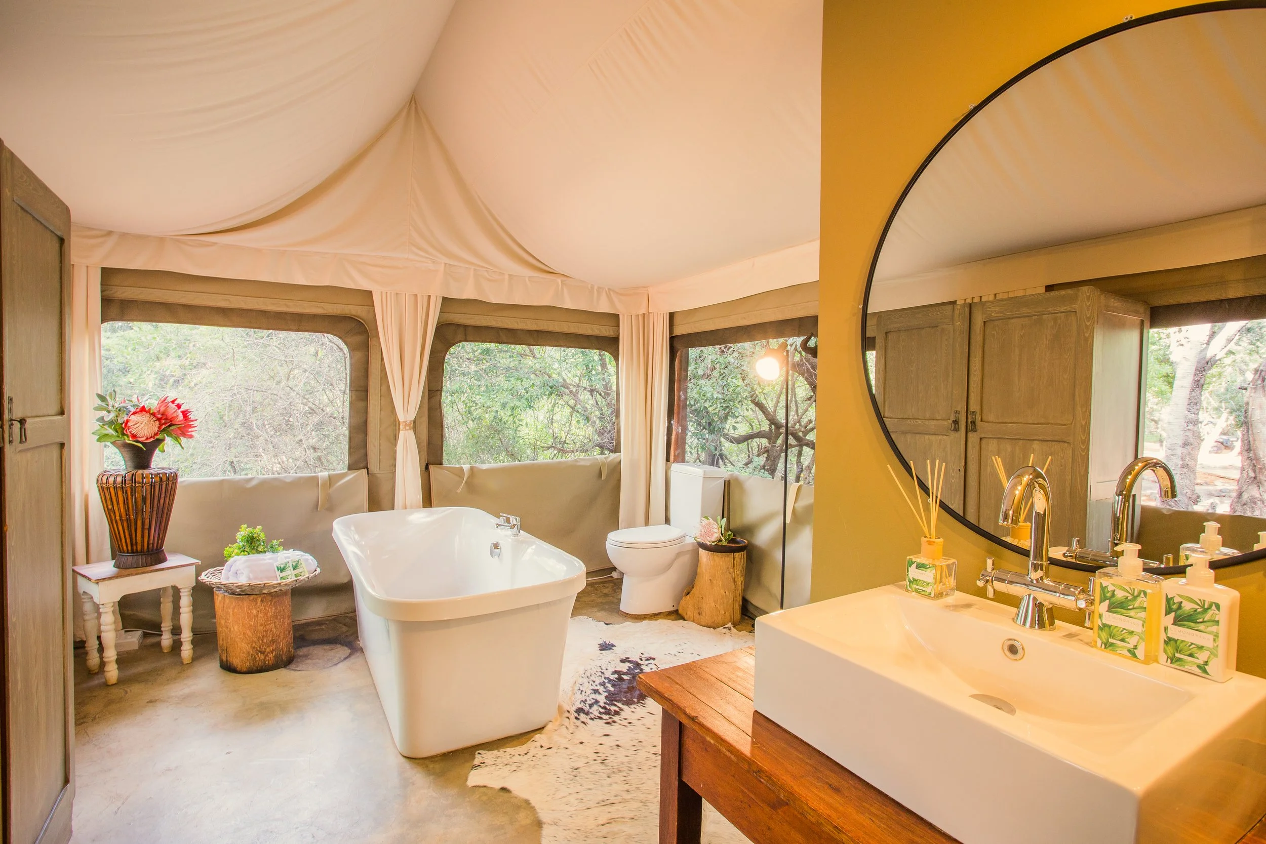 Bathroom with freestanding bathtub, toilet, vanity with round mirror, and large windows with view of trees.