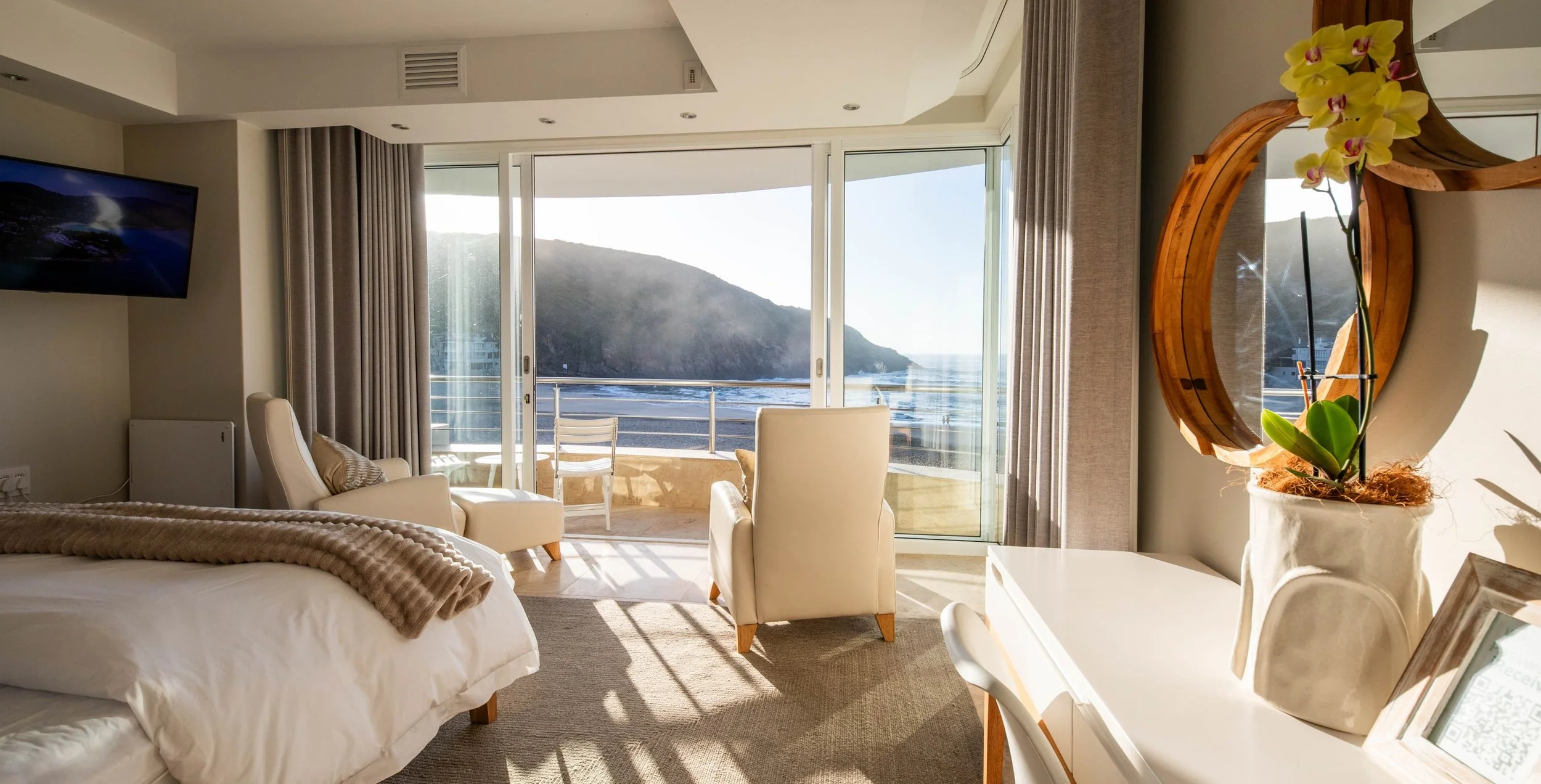 Bright hotel room with sliding glass doors leading to a balcony overlooking the ocean and a mountainous landscape. The room features beige and white furniture, including a bed with a beige throw, a white armchair, a white desk with a framed photo, and a large flowerpot with a yellow orchid. There is a wall-mounted flat-screen TV to the left.