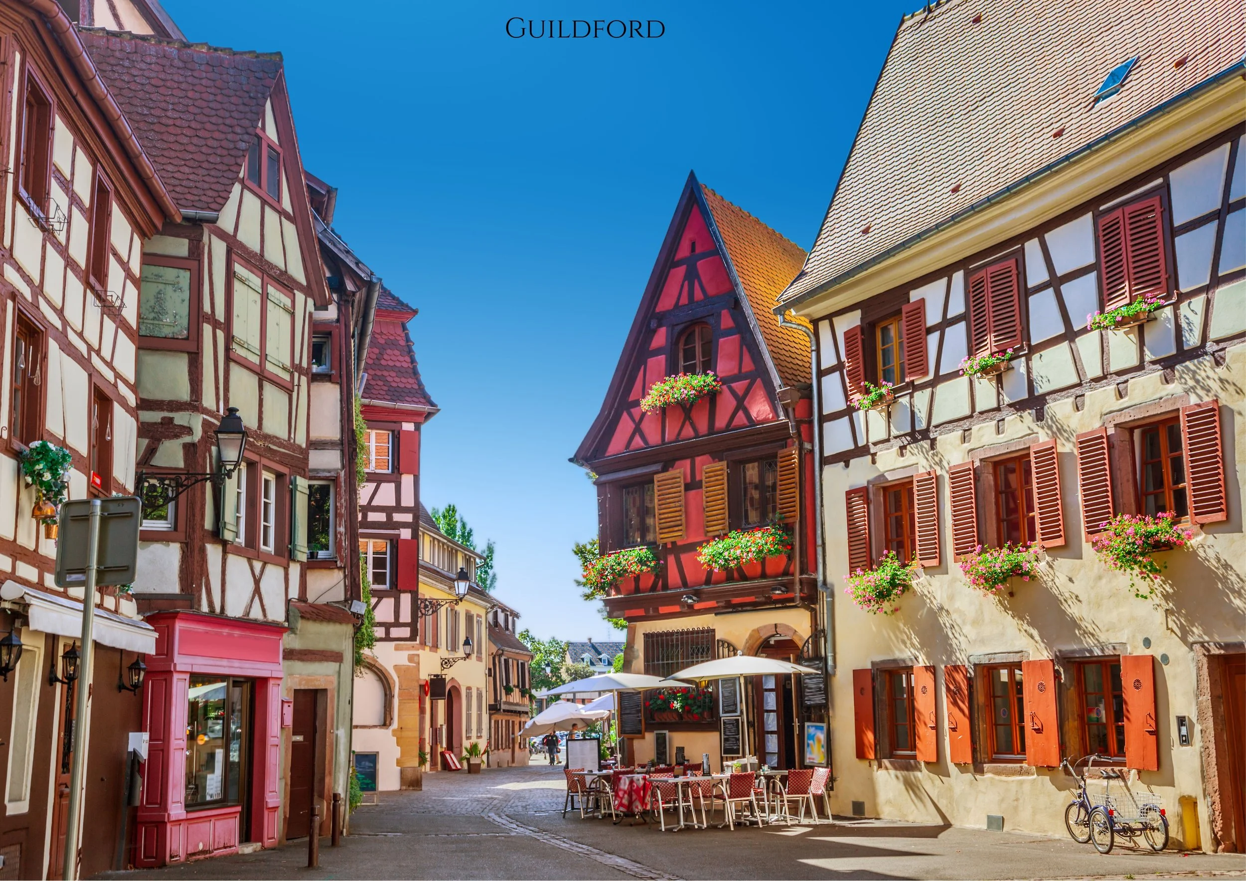 A picturesque European street scene with traditional half-timbered houses, colorful facades, flower boxes on windows, outdoor seating with tables and umbrellas, and a clear blue sky.