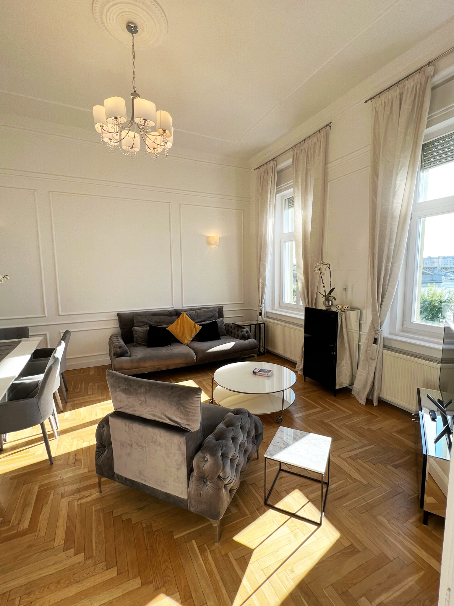 Living room with a velvet tufted armchair, a gray sofa with a yellow pillow and black pillows, a round white coffee table, and large windows with white curtains. Overhead chandelier and sunlight on wooden parquet flooring.