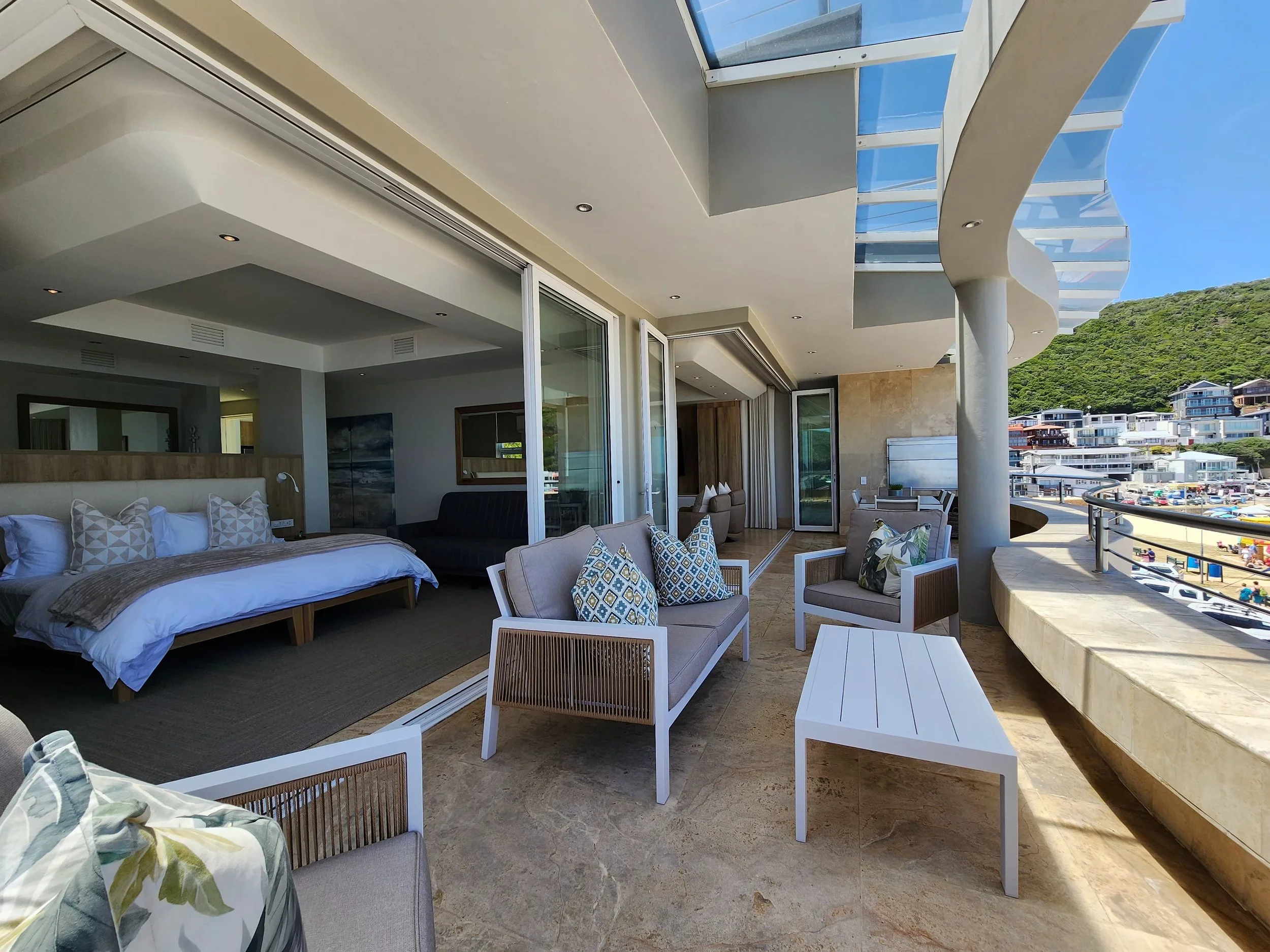 A spacious balcony with outdoor seating, a view of a beach and hillside with houses, and part of a bedroom inside the house.