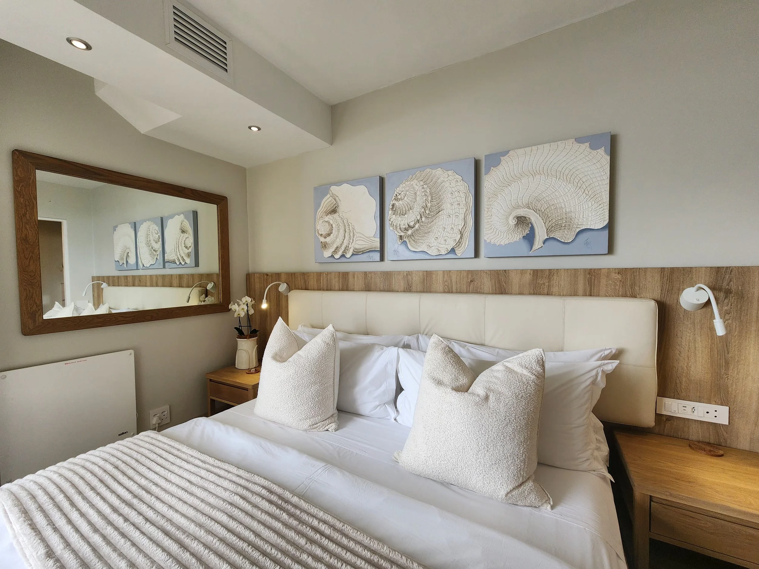Bedroom with a bed, white pillows, and beige decorative cushions, a wooden headboard with artwork of seashells, a large mirror, and side tables with lamps