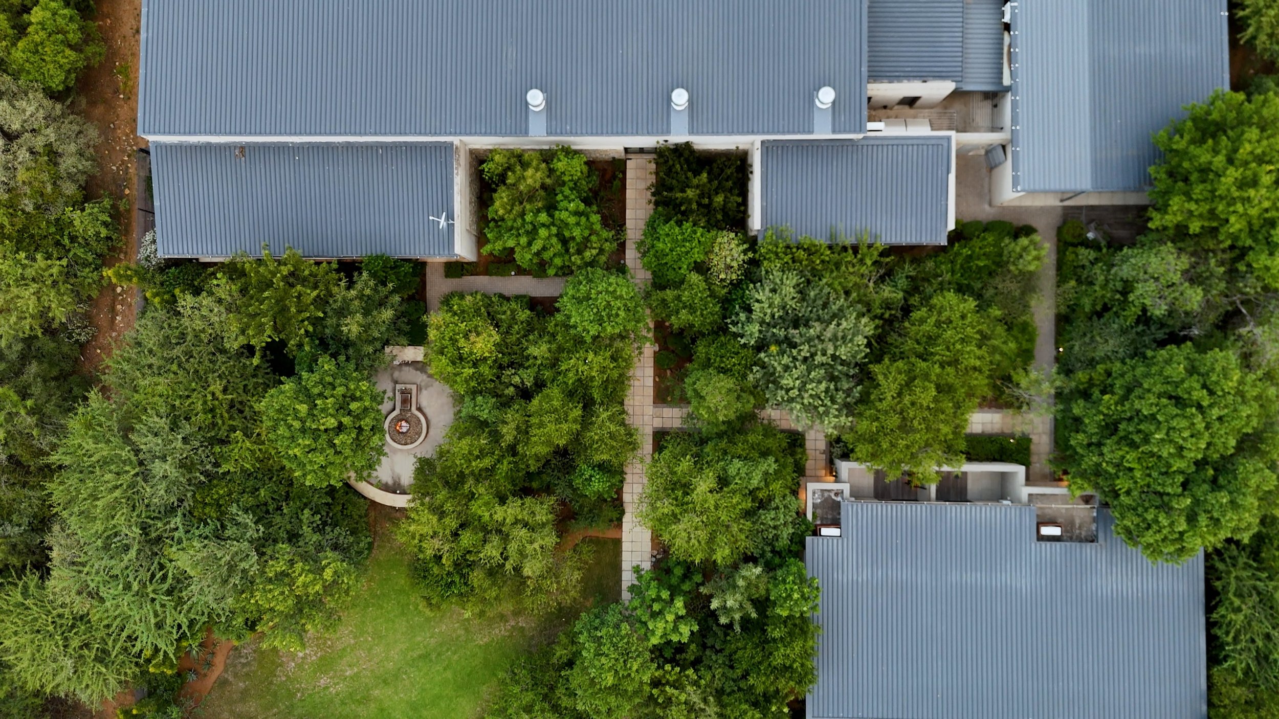 Top-down aerial view of a residential area with several buildings, gray metal roofs, a garden with lush green trees, pathways, and a small circular structure with a fire pit or fireplace.
