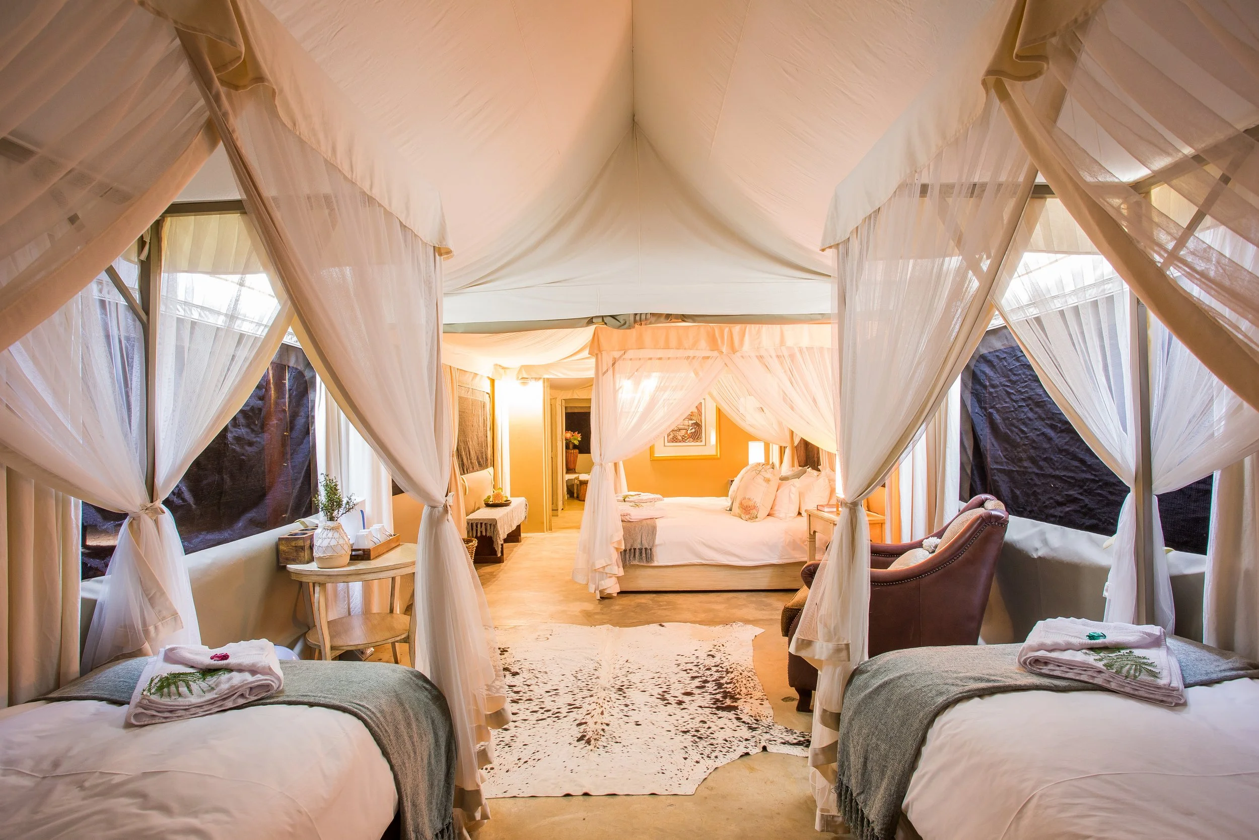 A luxurious bedroom with two beds with draped white canopies, a sitting area with a brown armchair, a rug on the wooden floor, and a large bed in the background with white linens and pillows.