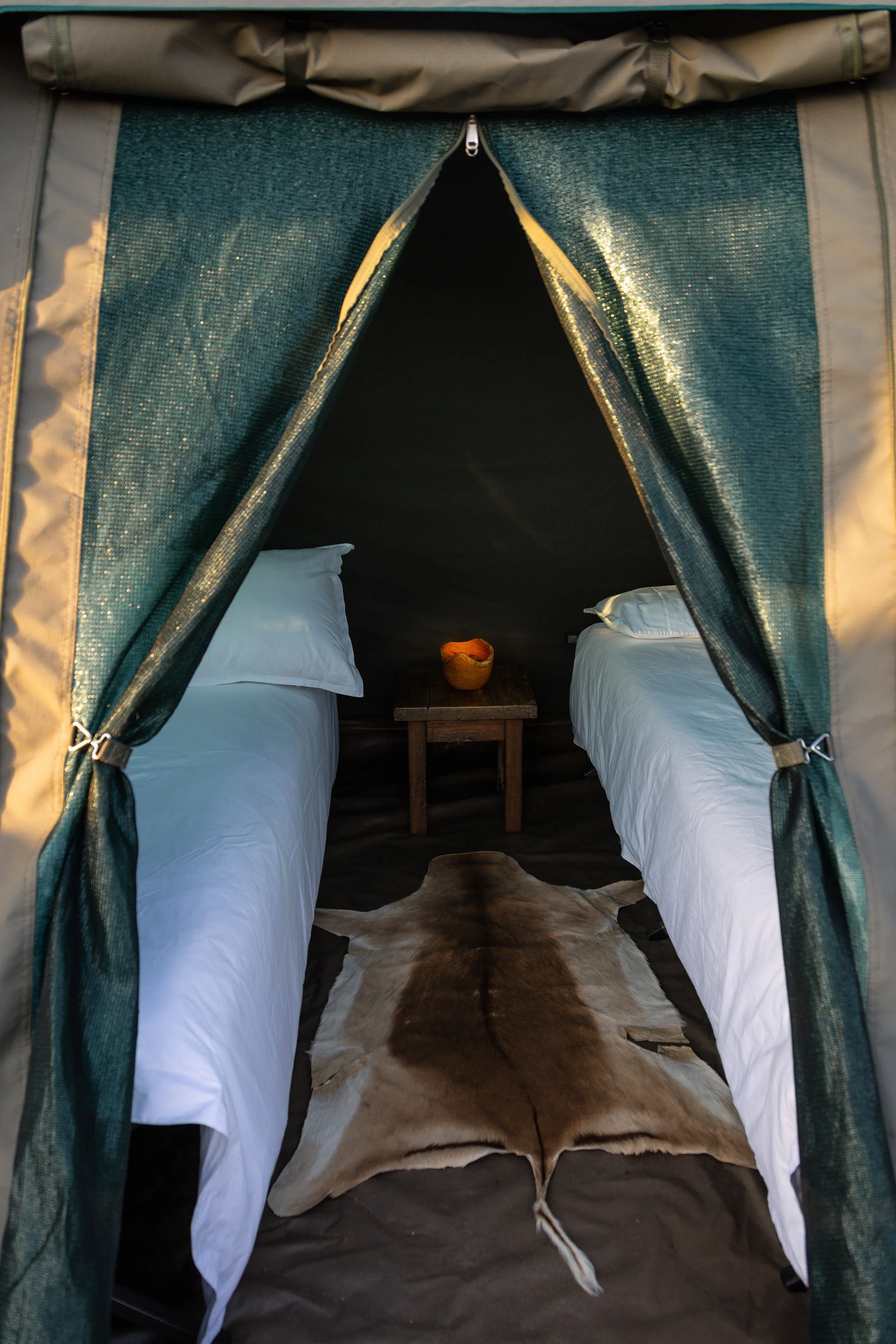 Interior of a camping tent with two beds, a small wooden table with a bowl between them, and a cowhide rug on the floor.