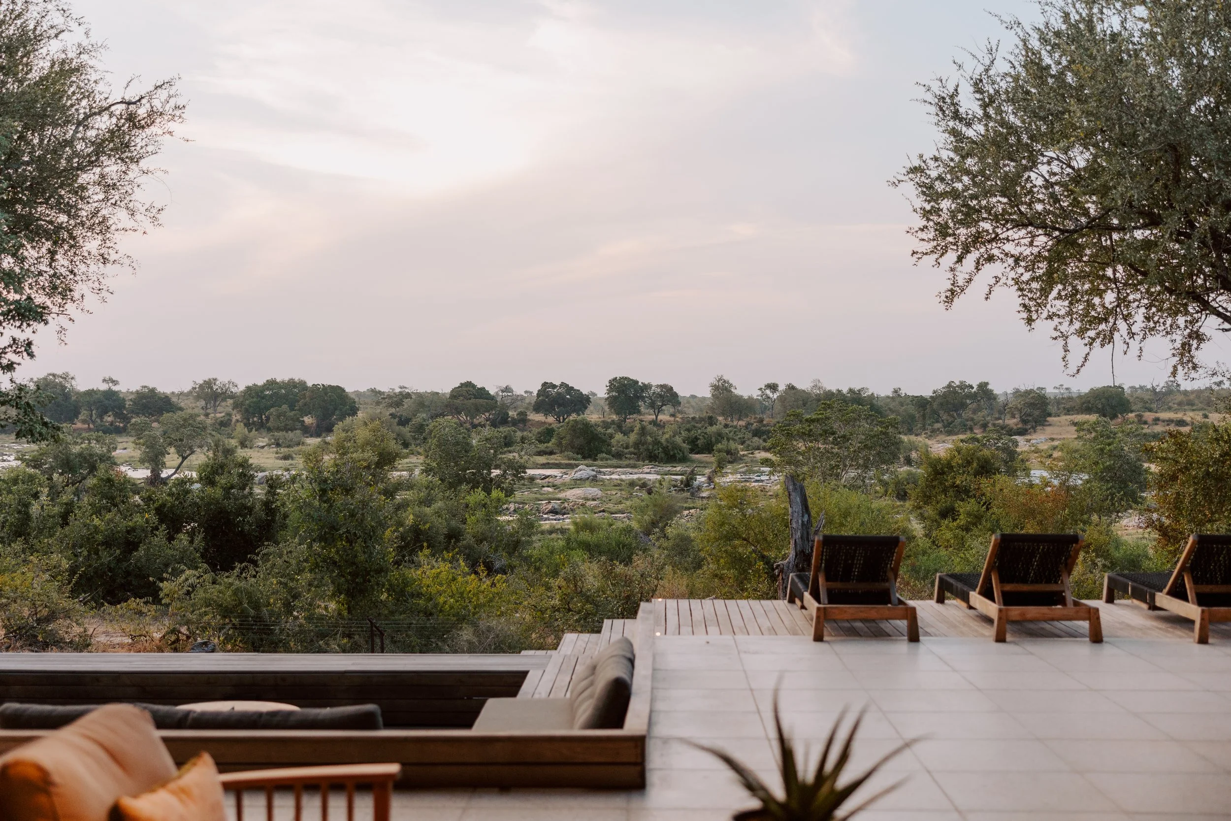 Outdoor patio with lounge chairs overlooking a lush, green savannah landscape with trees and a river in the distance, under a cloudy sky.