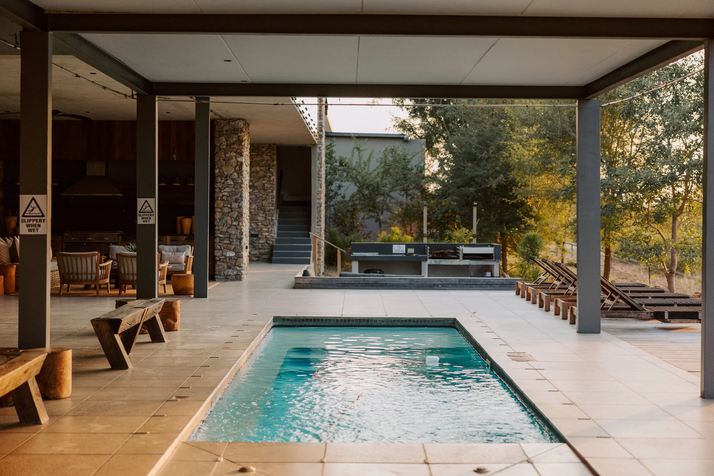 Outdoor pool area with lounge chairs, a barbecue grill, and a shaded seating area with wooden benches and chairs, surrounded by trees and greenery.